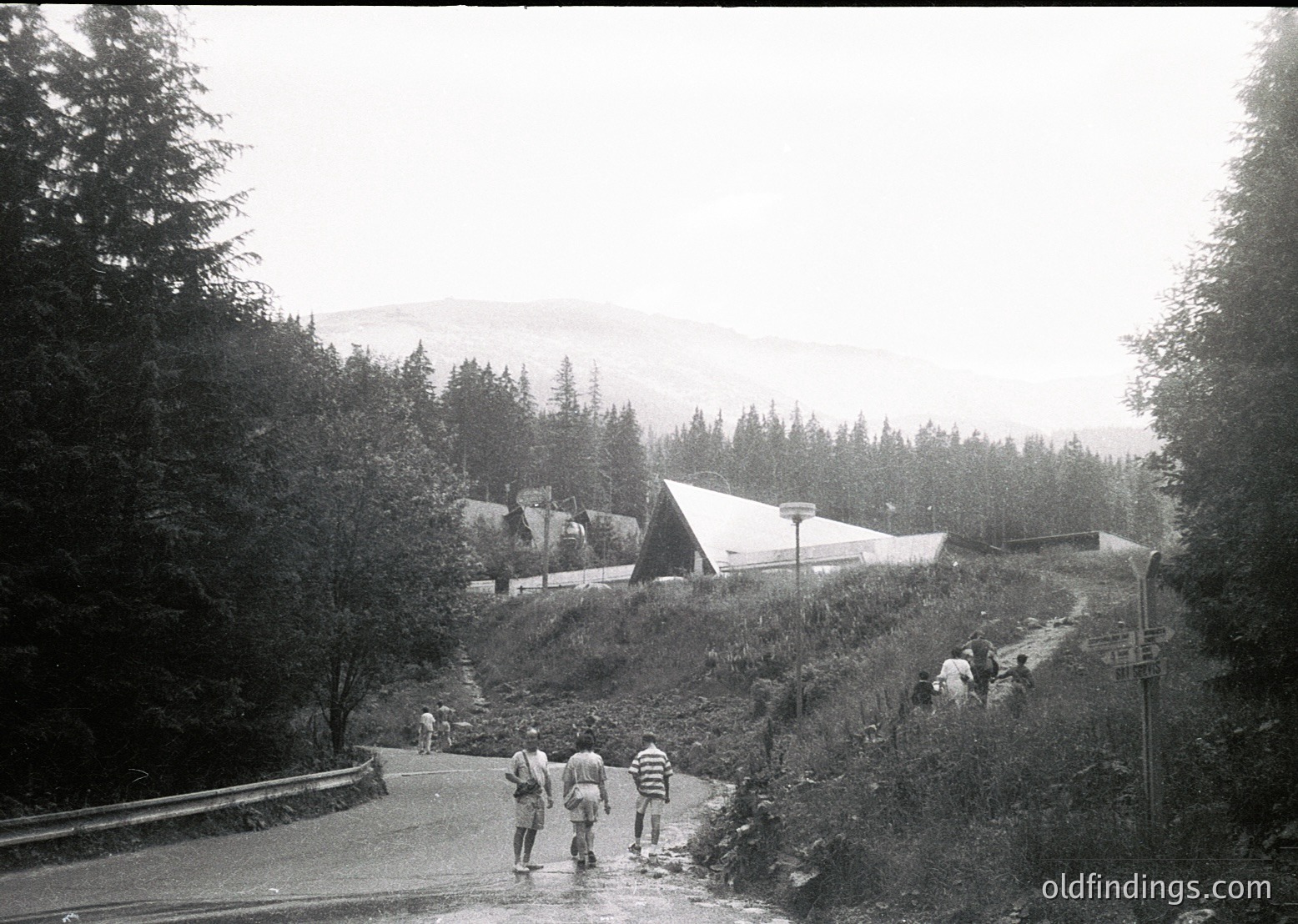 Black-and-white alpine roadside scene with dense coniferous forest and misty mountains in background. Three hikers in mid-20th-century attire cross a shallow stream on a winding road, flanked by grassy slopes. A small wooden cabin and utility pole suggest remote mountain settlement.