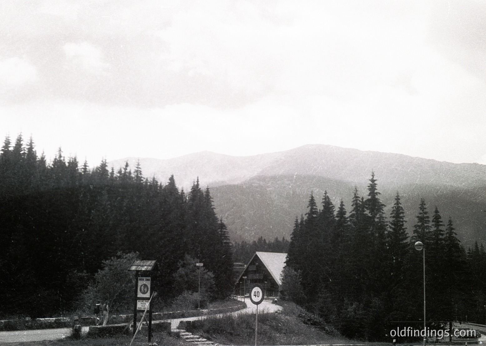 Black-and-white alpine roadside scene featuring a small wooden chalet-style building with a circular sign displaying "101" and a road sign indicating a 10 km/h speed limit. Dense coniferous forest surrounds the area, leading to misty, layered mountain peaks in the background. Likely a European mountain pass or resort entrance, mid-20th century.