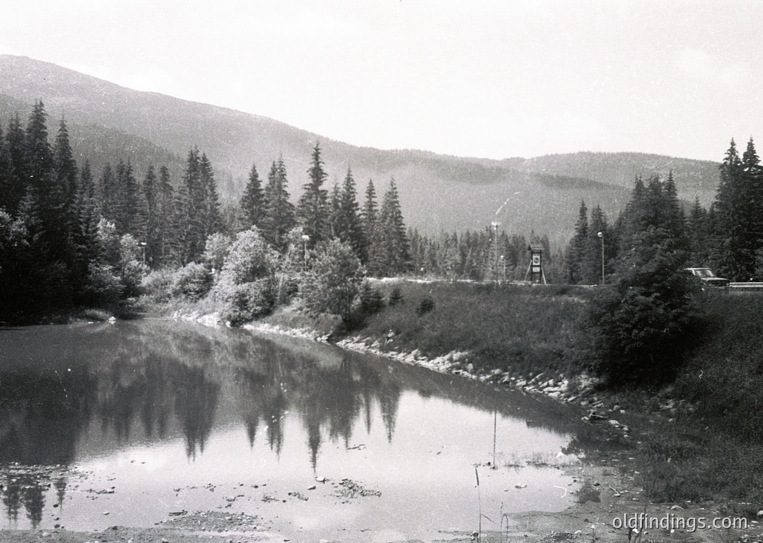 Mid-20th century black-and-white alpine scene: serene river winding through forested valley with snow-capped peaks in background. Small wooden bridge and utility tower near right bank. Dense coniferous trees frame reflective water surface.