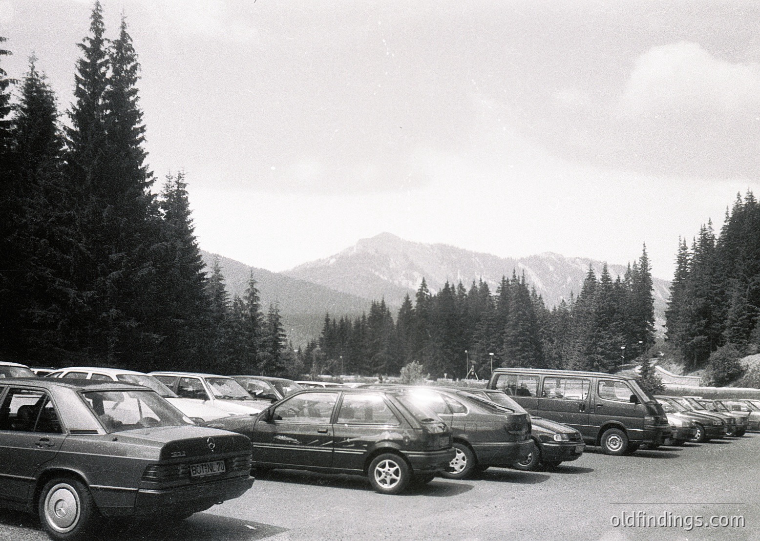 Black-and-white shot of a mountain parking lot with 1980s-era sedans and station wagons, likely European. Snow-capped peaks and dense coniferous forest frame the scene. Overcast sky suggests alpine weather.