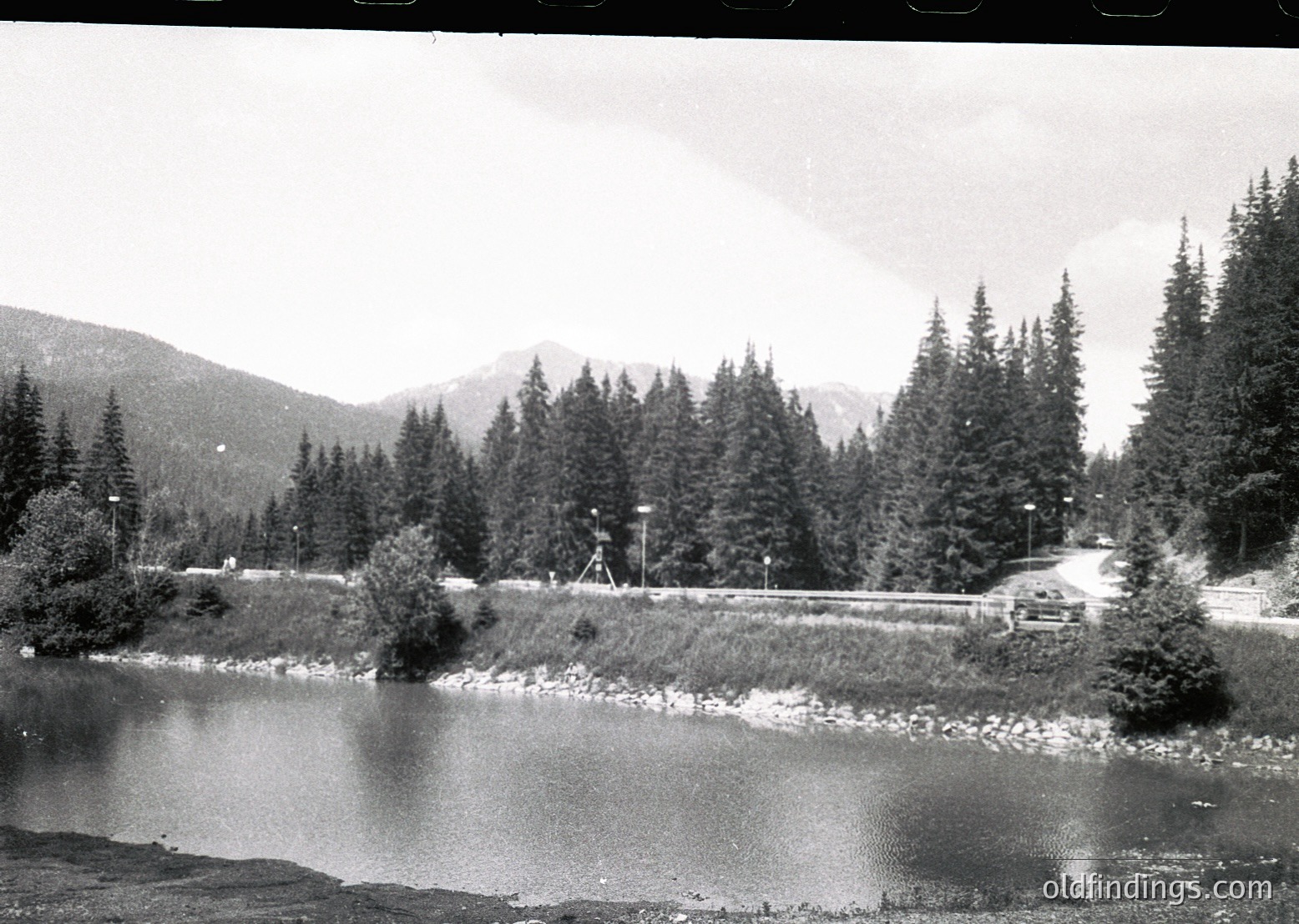 Mid-20th century black-and-white shot of a winding road alongside a serene alpine lake, framed by dense coniferous forests and snow-capped peaks. A single car travels along the road, while utility poles line the path.