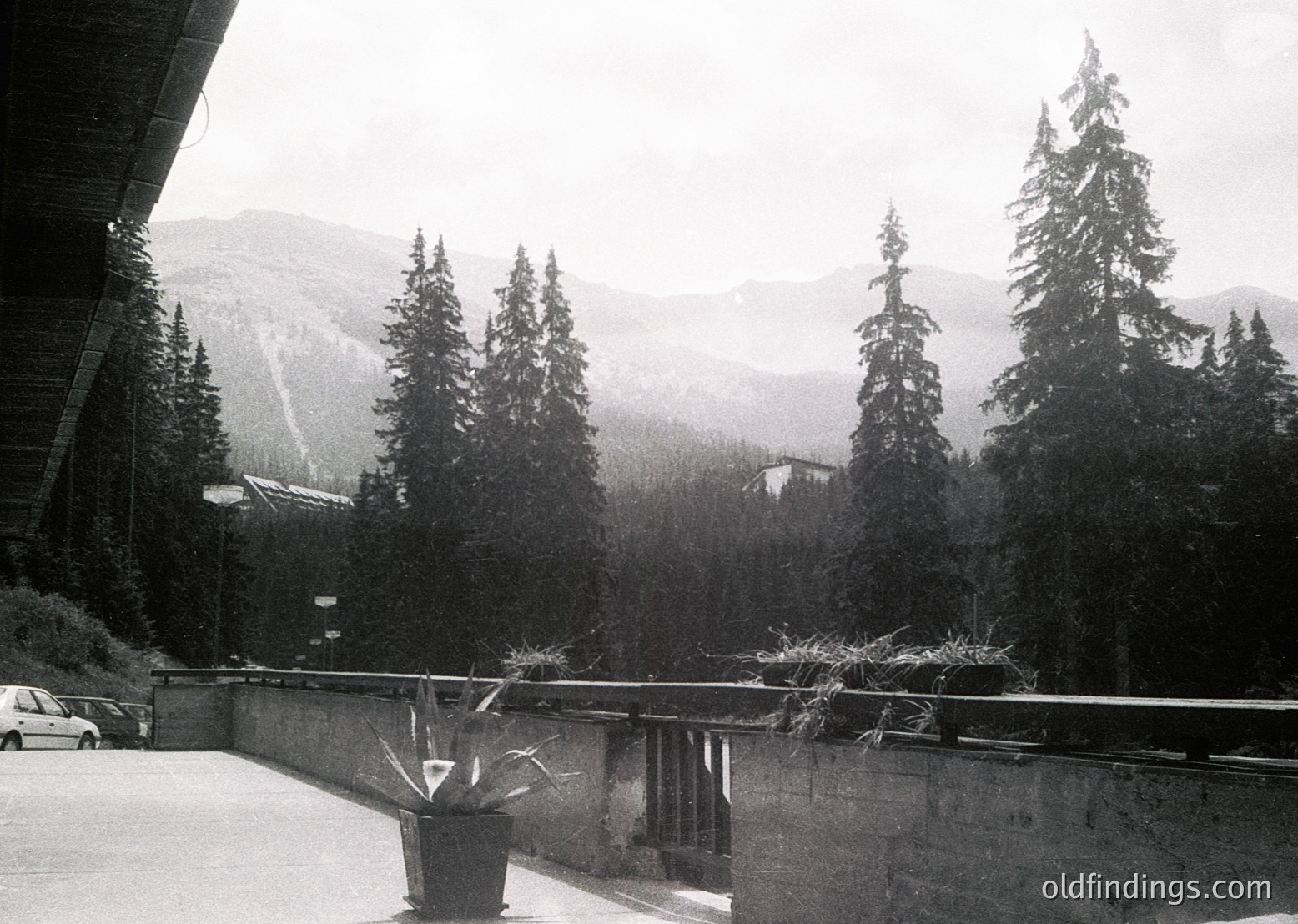 Black-and-white alpine resort scene featuring modernist concrete balcony with metal planters. Snow-capped peaks and dense coniferous forest frame the horizon. Mid-century architectural style suggests a 1960s-70s ski lodge or mountain retreat.