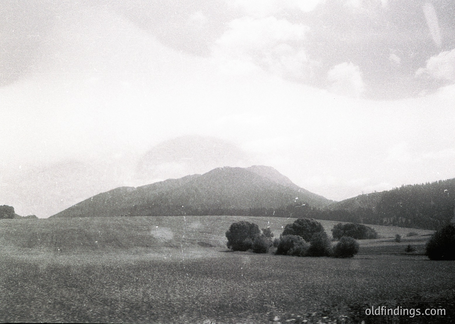 Vintage black-and-white landscape featuring rolling hills, sparse trees, and misty mountain peaks under diffused light. Mid-20th century aesthetic with soft focus and high-contrast tones.