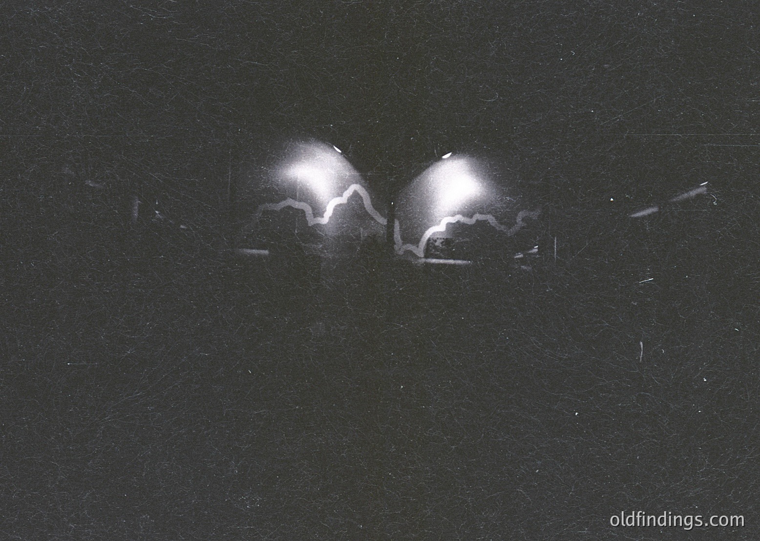 High-contrast black-and-white shot of two symmetrical, illuminated headlights against a dark background, likely from a vintage or classic car. The sharp, focused beams suggest a nighttime setting. Ideal for automotive design references or historical vehicle documentation.