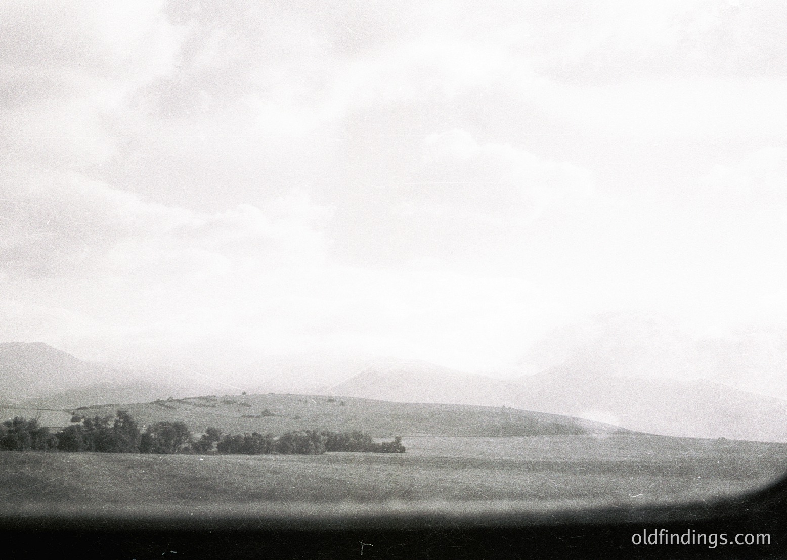 Vintage black-and-white landscape featuring rolling hills, sparse tree clusters, and misty horizon under overcast skies. Evokes mid-20th century rural scenery, likely European.