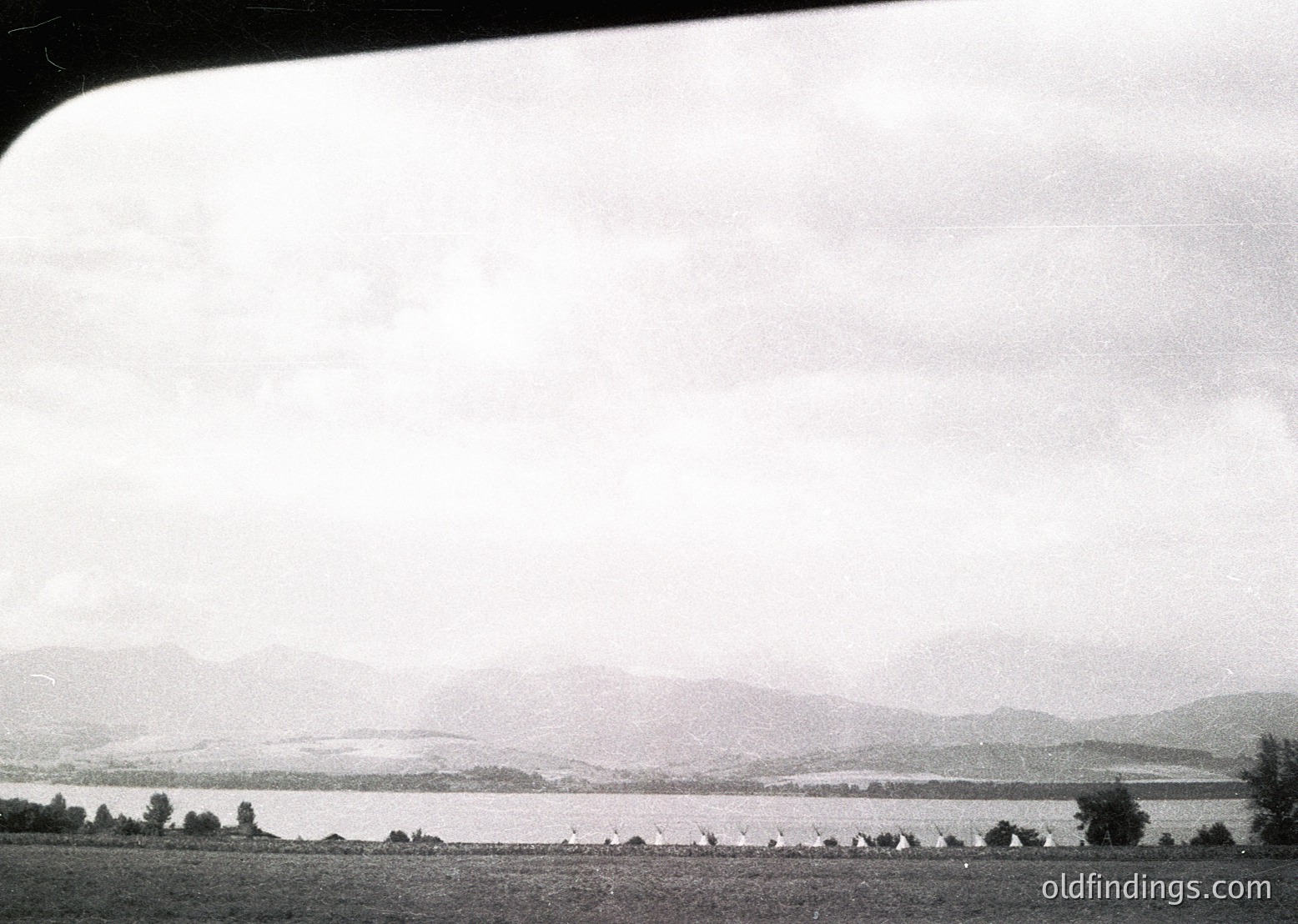 Mid-20th century black-and-white aerial view of a coastal plain bordered by low hills. Dense tree line frames a narrow strip of land leading to a body of water, likely a lake or sea inlet. Minimal human activity visible—sparse structures and pathways suggest rural or undeveloped area.