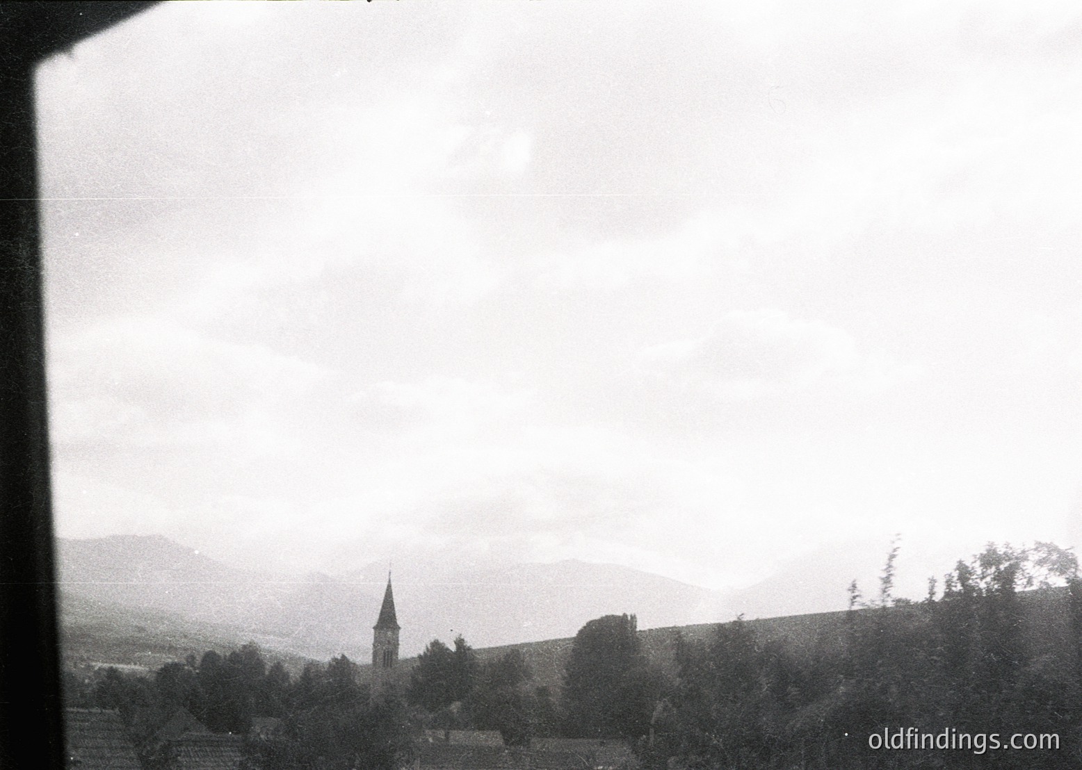 Vintage black-and-white landscape featuring a lone church steeple rising above rolling hills and dense forest. Mist obscures distant mountain peaks, enhancing the serene, timeless atmosphere. Likely European countryside, mid-20th century.