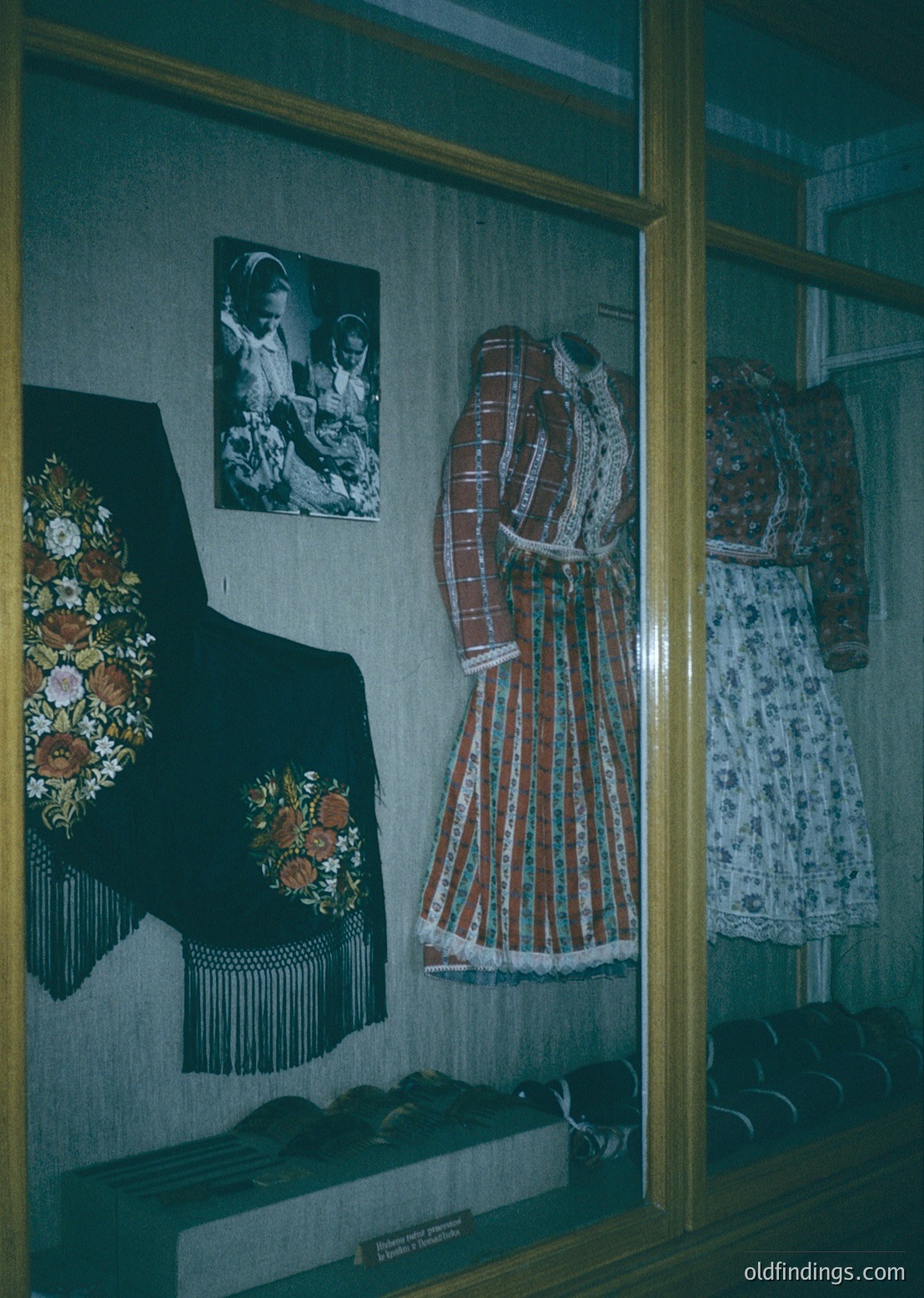 Traditional folk costumes displayed in a museum vitrine, featuring embroidered vests, striped skirts, and headpieces with floral motifs. Black-and-white photos of women in similar attire flank the display. Likely Eastern European, possibly Bulgarian or Romanian, 20th-century folk heritage.
