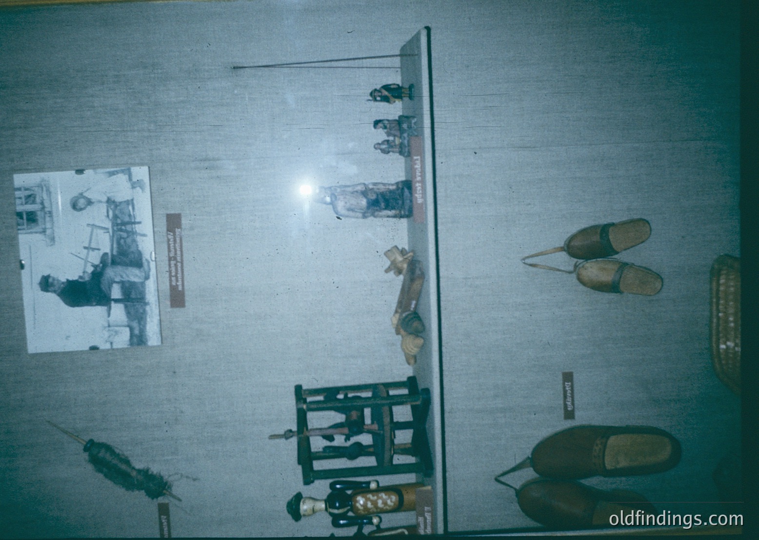 Vintage display case showcasing historical footwear and tools, likely from a museum or archive. Includes wooden clogs, a leather boot, and a vintage shoe with visible wear. Adjacent to footwear: a blacksmith’s hammer, anvil fragment, and a rusted metal object. Black-and-white photo inset depicts a 19th-century blacksmith at work. Suggests industrial heritage, likely