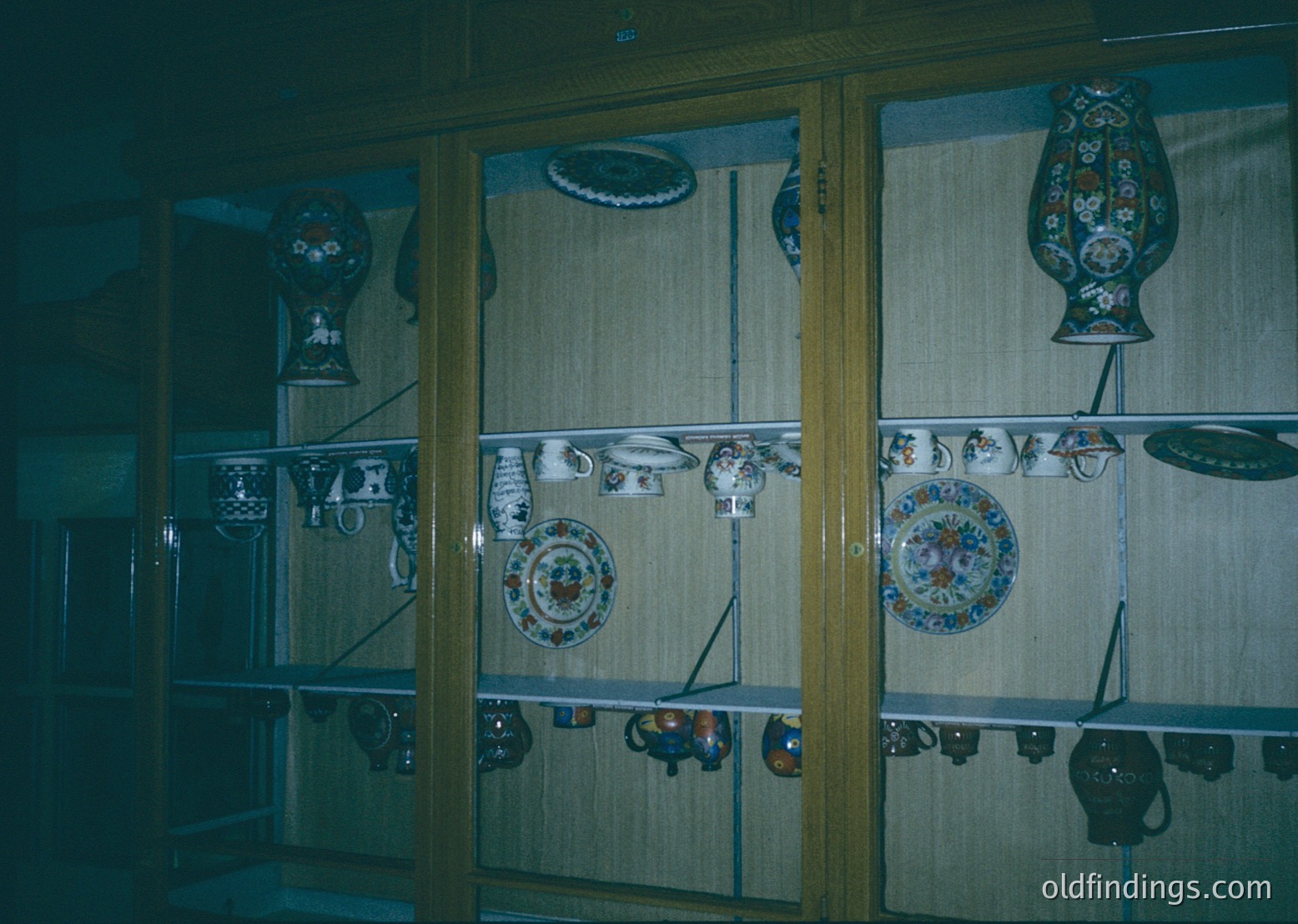 Vintage ceramic display showcasing ornate plates, pitchers, and teapots with floral/geometric patterns. Glass-front cabinet suggests museum or antique shop setting. Likely Eastern European folk art, 20th century.