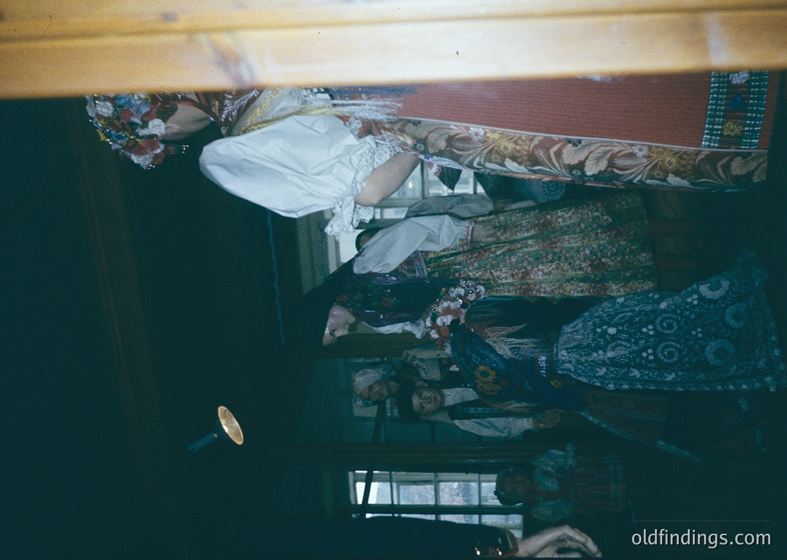 Vintage train compartment with two passengers seated in bunk beds. Upper bunk features floral-patterned fabric and a white pillow; lower bunk shows patterned textiles and a brass-colored handle. Reflections in the window reveal a blurred landscape. Likely mid-20th century European rail travel.