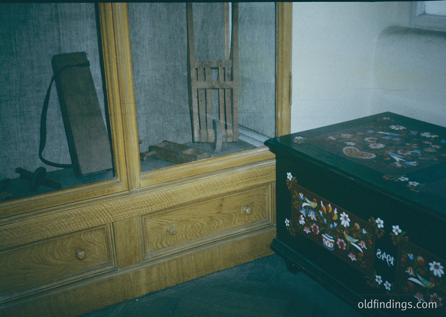 Vintage wooden furniture featuring a dark-stained dresser with brass handles and a floral-patterned trunk. Reflective mirror reveals a ladder and rustic tools. Likely Eastern European, 19th–early 20th century.