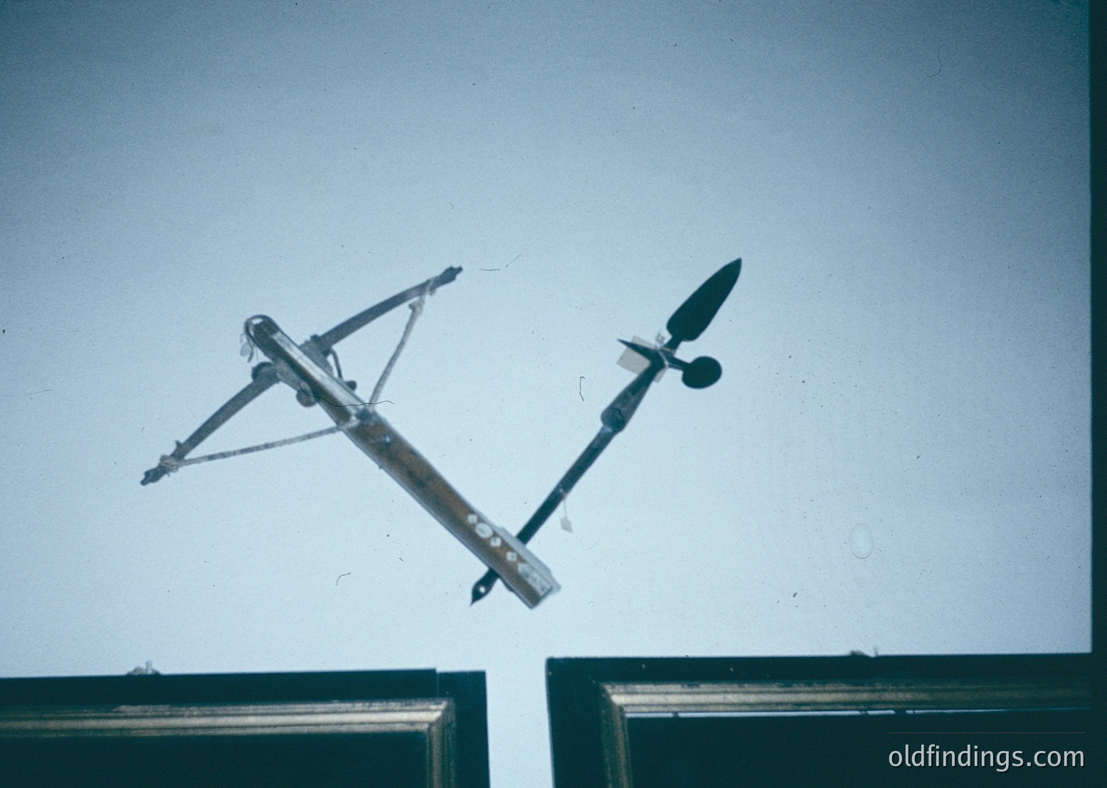 Vintage wind vane mounted on a metal frame, likely from mid-20th century industrial or agricultural site. The design features a horizontal crossbar with a vertical tail fin and directional arrow.