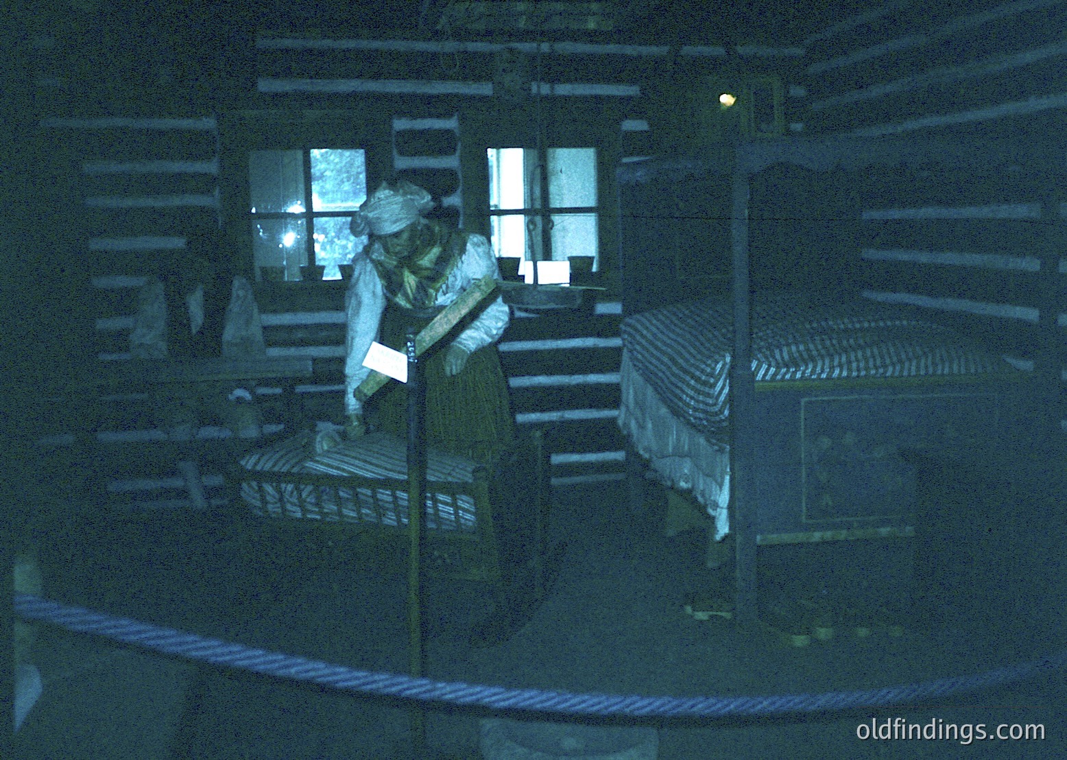 Vintage interior of a rustic wooden cabin, likely late 19th to early 20th century. A woman in period attire—apron, blouse, and headscarf—stands near a bed with striped bedding, holding a book or paper. Wooden beams, a small window with glass panes, and a simple wooden table with a pitcher complete the scene. Evokes domestic life in rural settings.