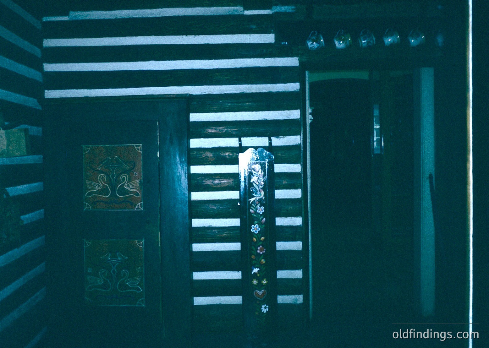 Dark, narrow wooden hallway with vertical slats, illuminated by a single dim light source. Decorative wall tiles featuring stylized floral motifs on left. Hanging garlands with artificial flowers and tassels on central door. Possible temple or shrine interior, suggesting cultural/religious significance.