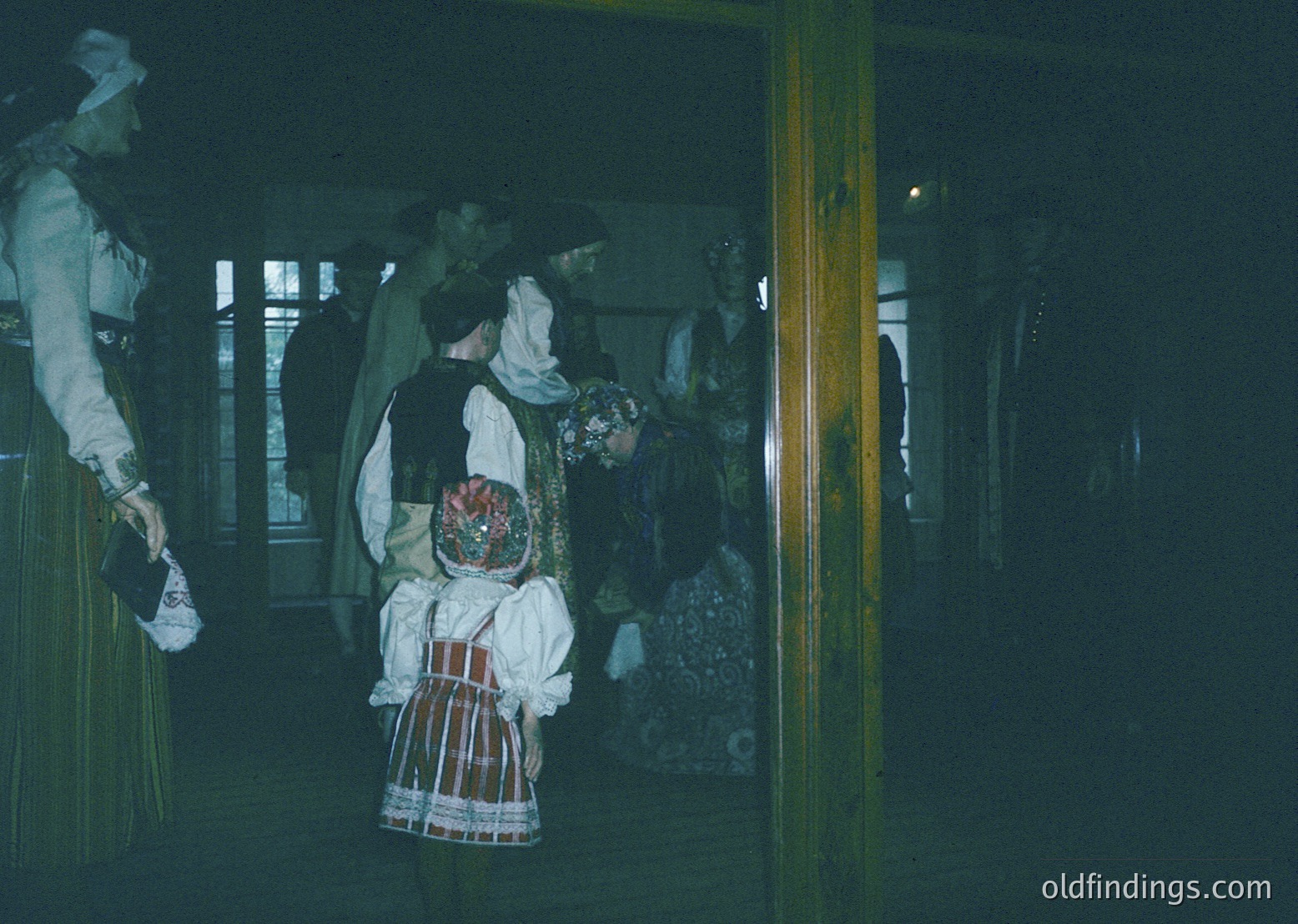 Traditional folk costumes in low-light indoor setting, likely a cultural event or festival. Intricate embroidered vests, striped skirts, and headscarves suggest Eastern European heritage. Wooden beams and dim lighting enhance rustic ambiance. Potential –1980s timeframe based on style.