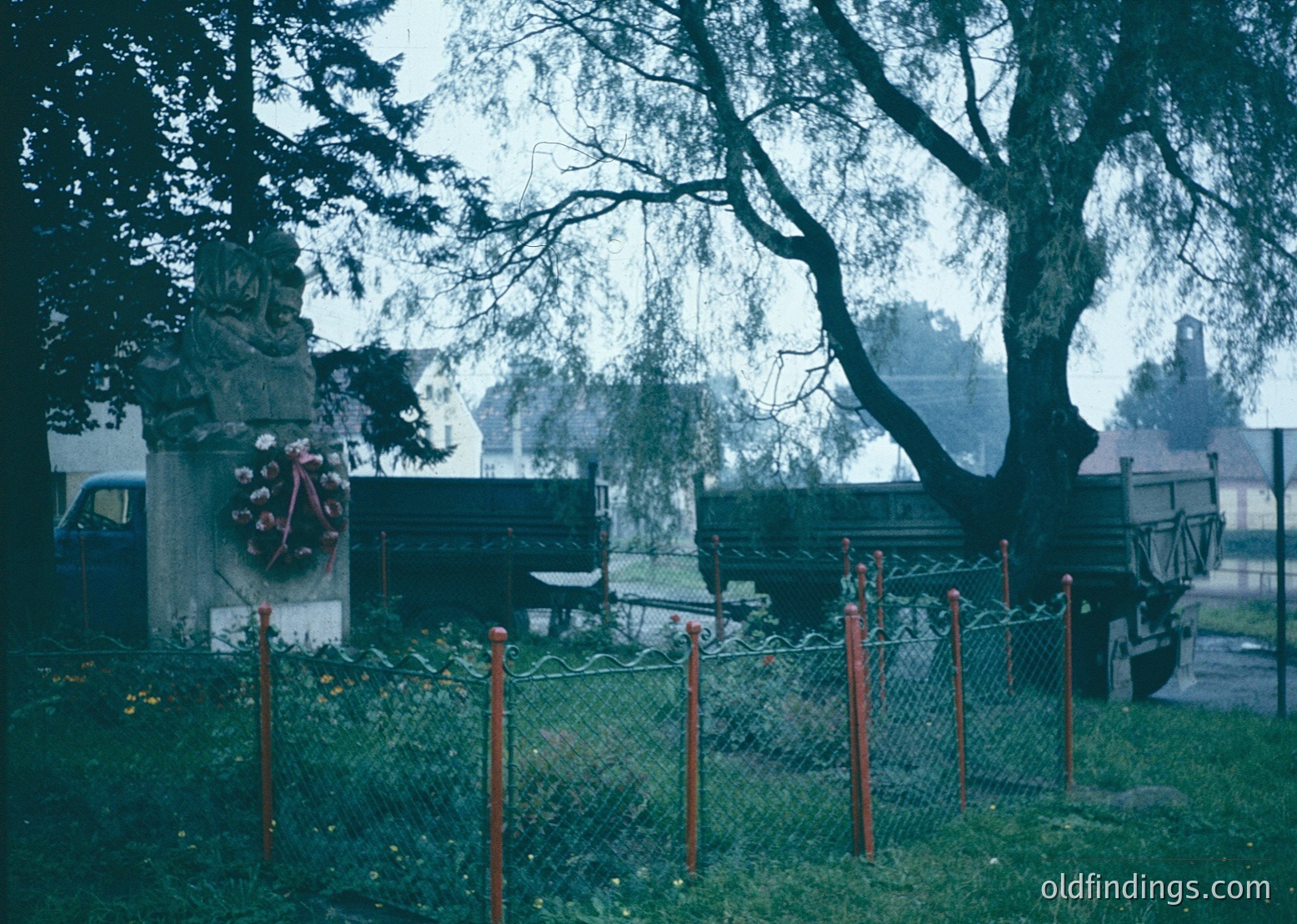 Vintage cemetery scene with Soviet-era monument featuring hammer-and-sickle emblem, surrounded by red-painted metal posts. Overgrown grass and parked vintage trucks in background suggest mid-20th century Eastern Bloc setting.