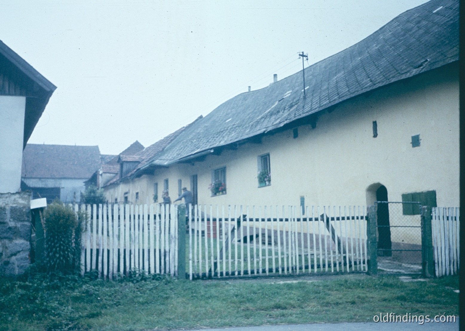 Vintage rural house with whitewashed walls and dark slate roofs, framed by a picket fence. Mid-20th century European farmstead, likely Eastern Bloc era. Simple wooden door and small window with floral curtains. Overcast sky suggests early photography era.
