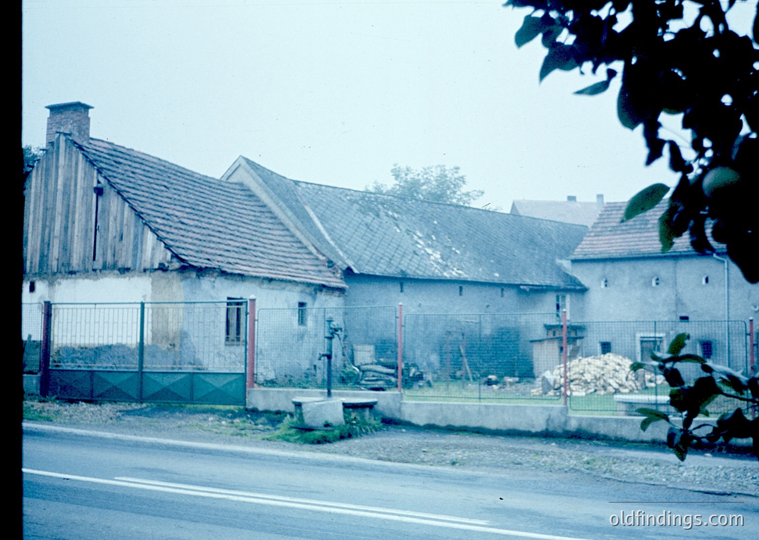 Vintage rural house with weathered brick and tiled roof, surrounded by chain-link fencing. Overgrown vegetation and construction debris in foreground. Likely Eastern European countryside, mid-20th century.