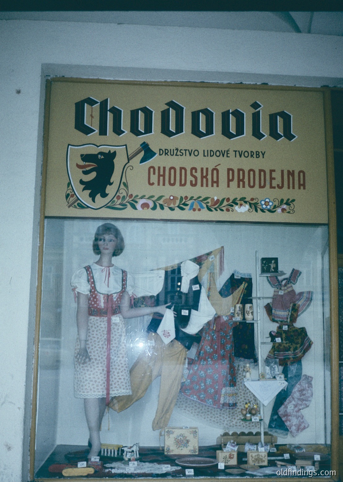 Vintage storefront sign for **Chodovin** (Czech: *Družstvo lidové tvorby Chodská prodejna*), a cooperative folk art shop from the 1960s–70s. Display features traditional embroidered textiles, ceramic items, and handcrafted folk costumes. Shield emblem with a bear symbolizes regional identity.