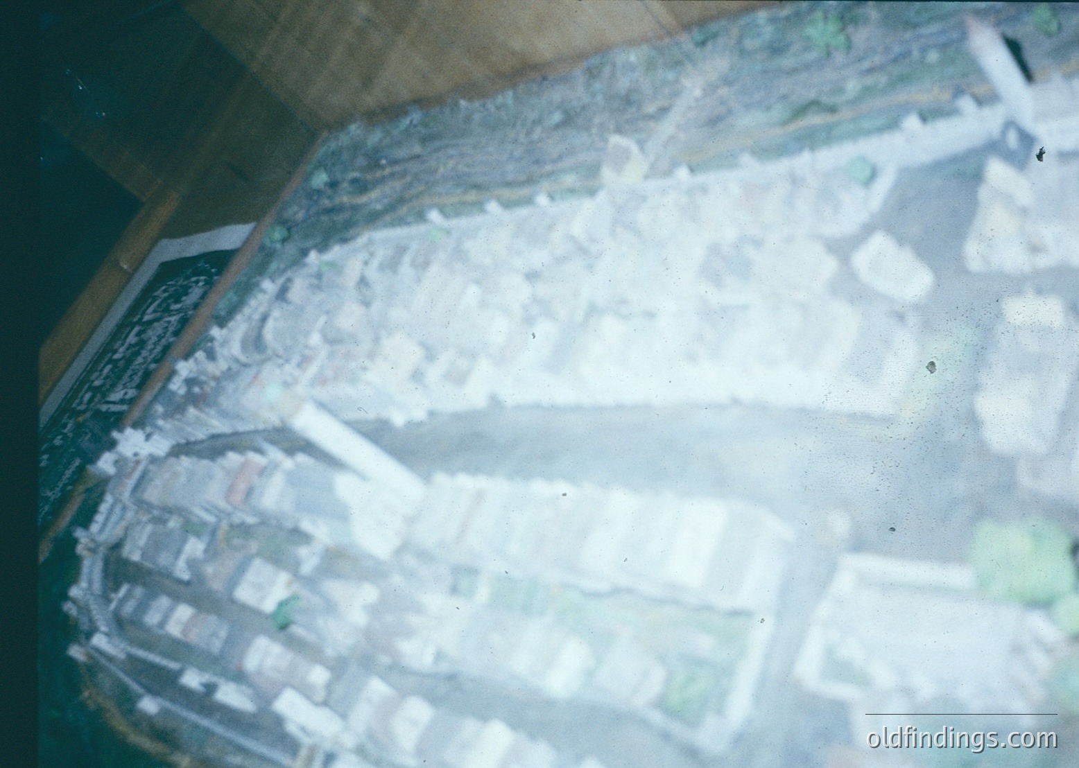 Aerial view of a coastal cityscape with dense urban layout, likely from the 1960s–1980s. Grid-pattern streets, low-rise buildings, and a prominent beachfront with light-colored sand dominate. Industrial or residential structures cluster near the water, suggesting port or harbor activity. Vintage film grain and sepia tone enhance nostalgic atmosphere.