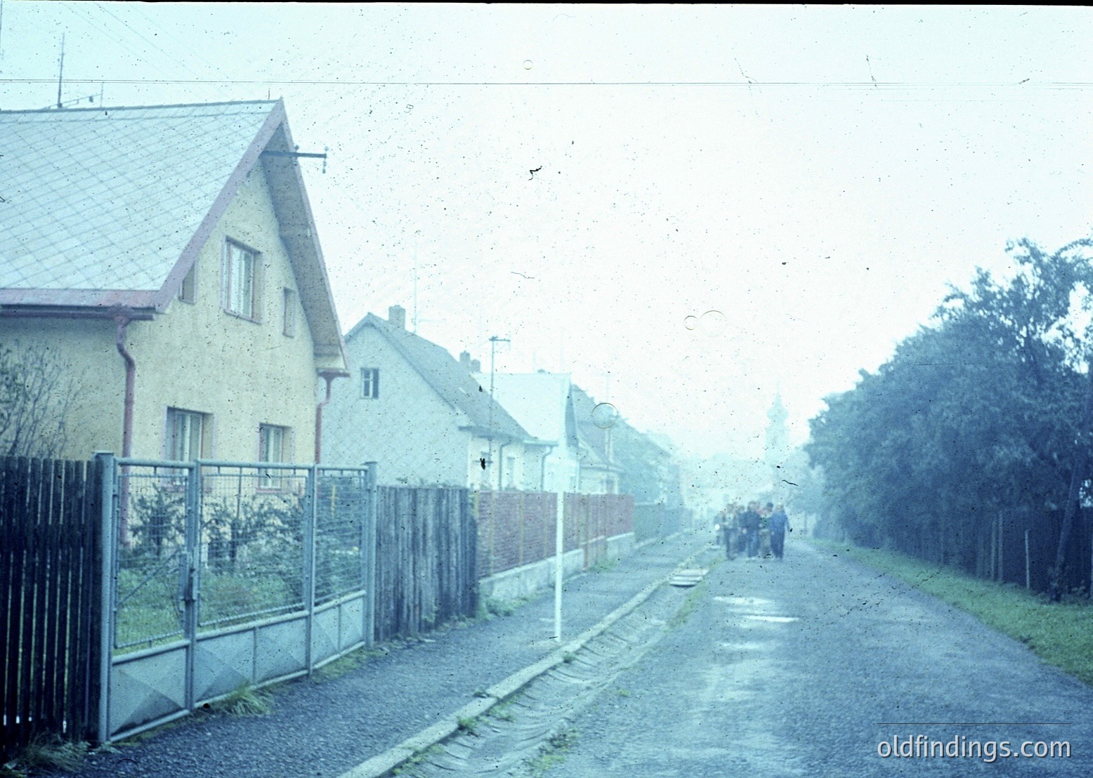 Vintage street scene featuring uniform Soviet-era apartment blocks with pitched roofs and chain-link fences. Foggy atmosphere suggests early morning or overcast conditions. Pedestrians in dark clothing walk along a wet, unpaved road flanked by trees and shrubs. Likely Eastern Bloc residential area, 1960s–1980s.