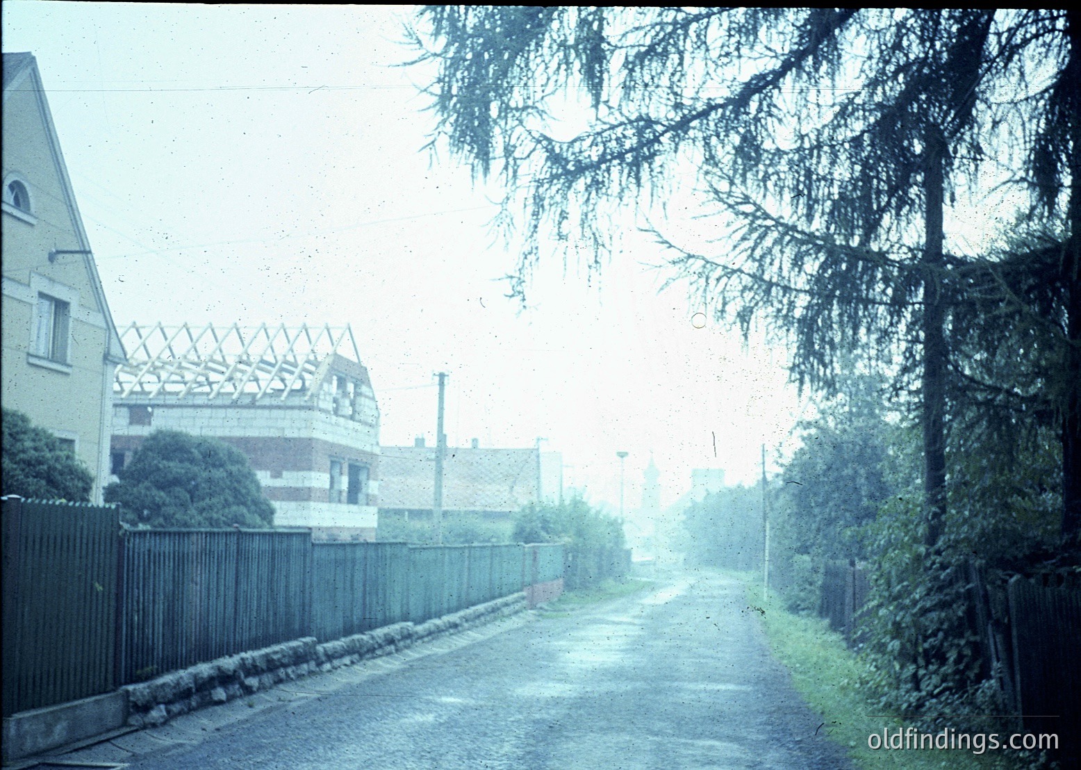 Vintage urban street scene with misty haze, featuring a narrow road flanked by stone curb and metal fence. Partially constructed concrete building under scaffolding in background, suggesting mid-construction era. Dense tree branches frame left side, adding depth. Likely 1960s-1970s industrial or transitional neighborhood.