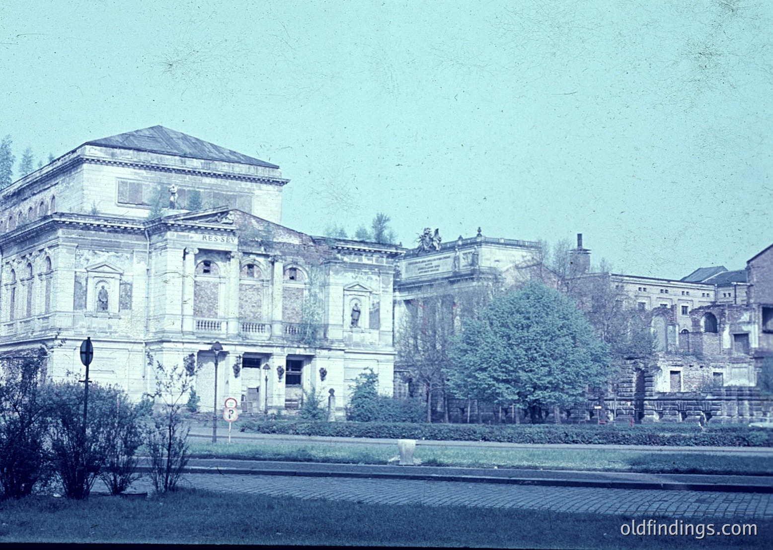Neoclassical building with symmetrical façade, arched windows, and decorative reliefs, likely from early 20th century. Courtyard and landscaped greenery in foreground. Urban street scene with vintage street lamp and road markings.