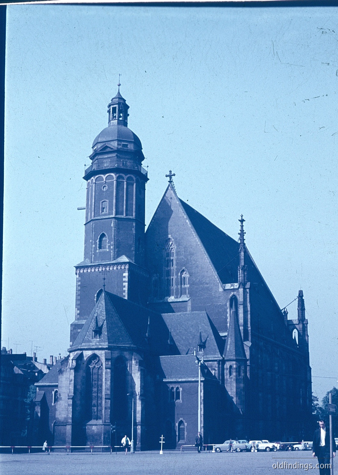 Historic Gothic Revival church with tall bell tower and pointed arches, likely 19th-century European architecture. Brick facade with decorative stonework and a steeply pitched roof. Mid-century street scene with vintage cars and pedestrians.