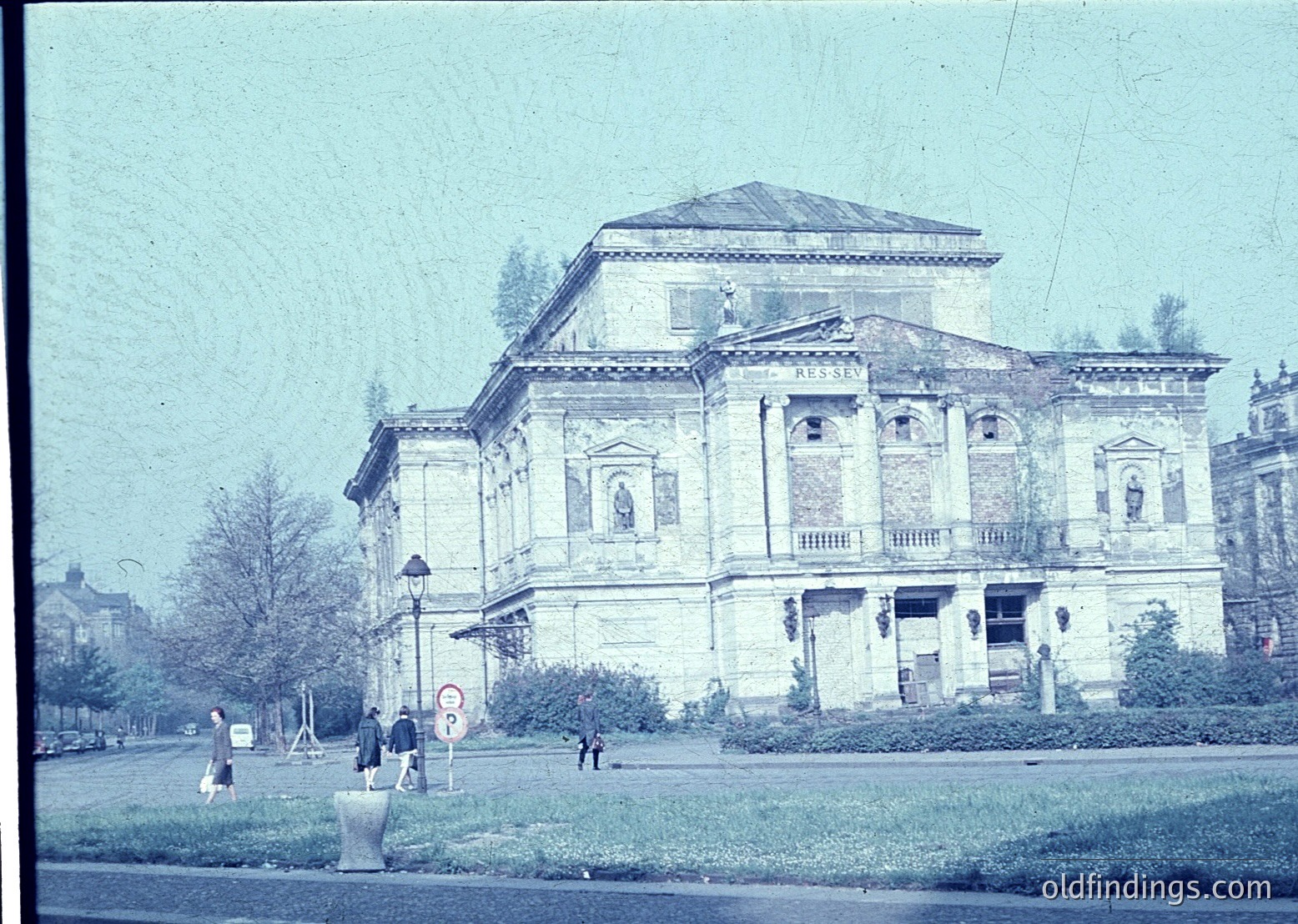 Neoclassical building with "RUSEV" inscription, likely a cultural or governmental structure from the early 20th century. Symmetrical facade features columns, arched windows, and a pediment. Pedestrians and vintage street lamp add urban context.
