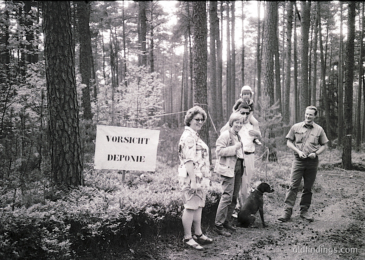 Family group photo in dense forest, 1960s Germany. Sign reads "VORSICHT DEPONIE" (Caution: Tip/Dump). Adults in mid-century attire, one holding child, another with dog.