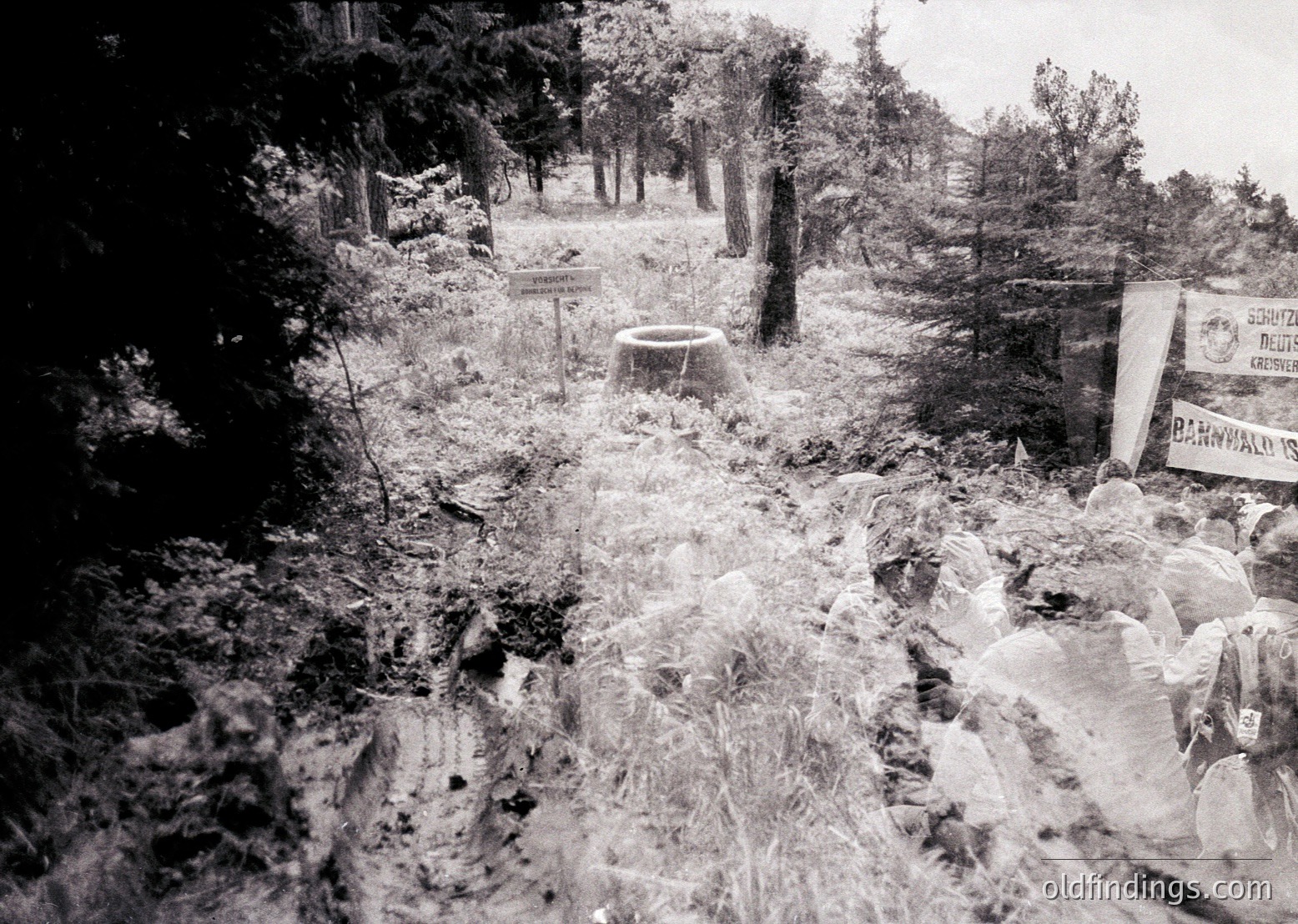 Snow-covered forest path with debris, likely post-war or post-disaster. Visible signs in Cyrillic suggest Eastern European location. Barrel and banner hint at wartime or occupation-era context.