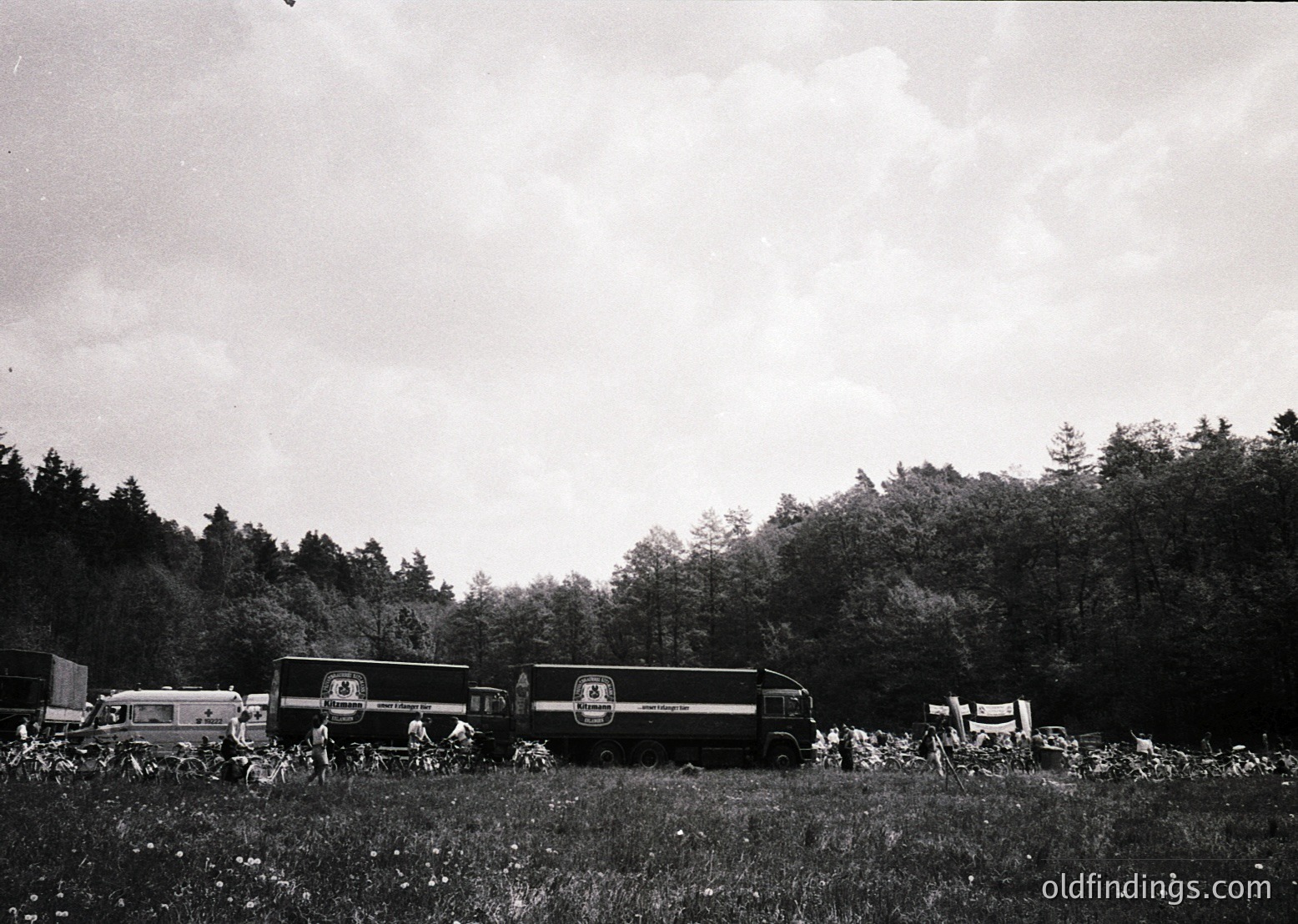 Vintage black-and-white photo of a vintage bus (likely 1950s–1960s) parked in a rural, wooded area with dense foliage. Group of people gathered around, some seated on grass, others standing near the bus. Log cabin-style structure visible in background.