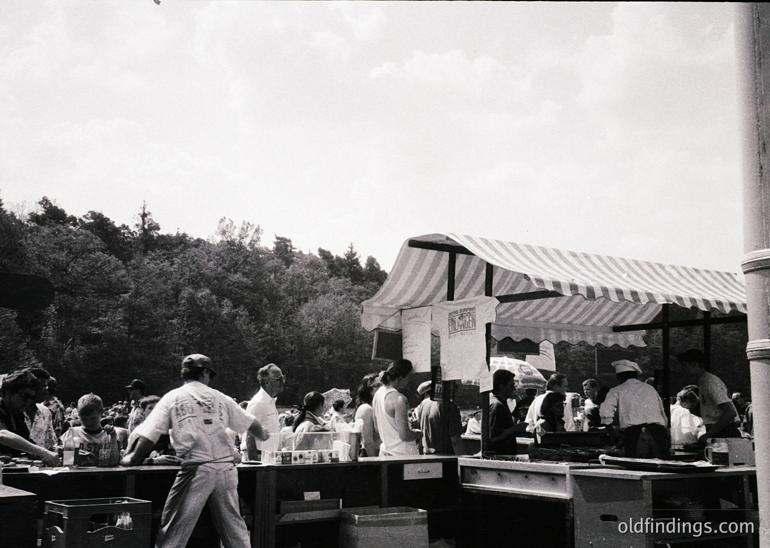 Black-and-white shot of a bustling outdoor food stall, likely from the **1950s–1960s**. Striped canopy with "MARSHMALLOW" signage suggests a dessert-focused vendor. Crowd in casual summer attire—shorts, hats, and aprons—indicates a communal event or fair. Wooden tables, metal coolers, and handwritten signs add to the vintage ambiance.