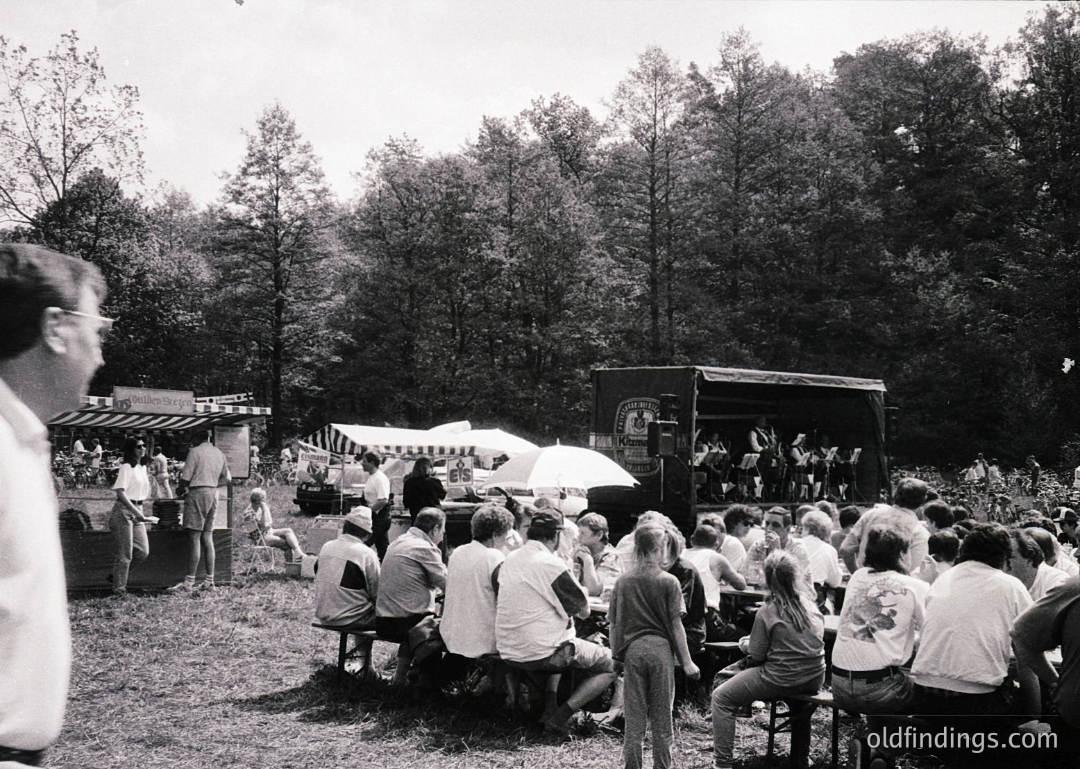 Outdoor concert or event in a wooded park setting, featuring a stage truck and seated audience on wooden benches. Mid-20th century attire suggests or . Crowd gathered under trees, indicating a natural, informal venue.