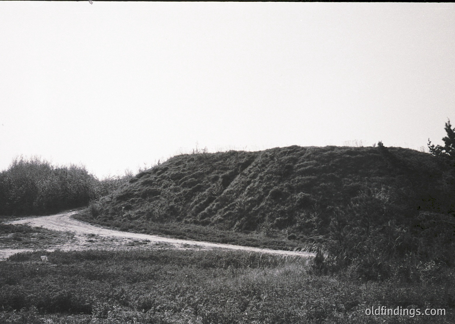 Dense, eroded earthen mound with layered, stepped terrain alongside a narrow, unpaved road. Vegetation includes sparse shrubs and grass. Likely a historic fortification or ancient burial site.