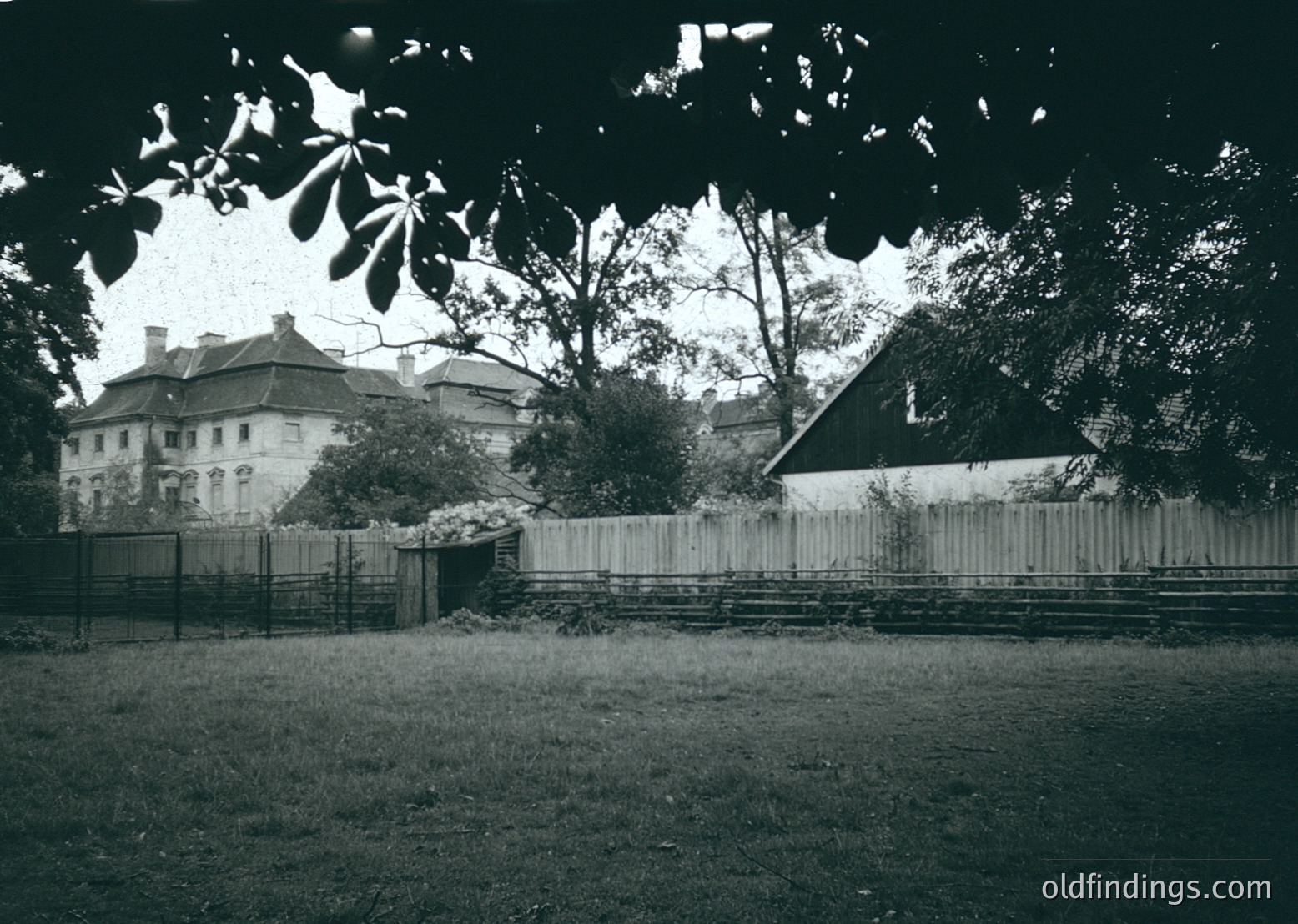 Vintage black-and-white photo of a European courtyard with a mix of residential and industrial elements. A two-story building with arched windows and a flat roof sits behind a weathered wooden fence. Adjacent is a sloped metal-roofed structure, likely a barn or workshop. Overgrown trees cast shadows across the scene, suggesting late afternoon light. The grassy area appears well-trodden, hinting at frequent use. Likely late 19th to early 20th century.