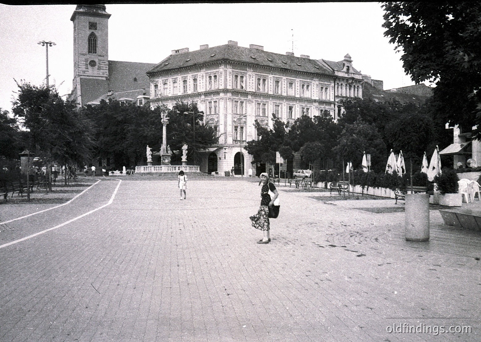 Mid-20th century urban plaza featuring a grand, symmetrical building with classical architecture—ornate façade, arched windows, and a clock tower. Two pedestrians, one in a patterned dress, walk along a wide, tree-lined road. Central monument with a statue and decorative lampposts. *(Note: The architectural style and monument suggest a European setting, likely Bulgaria based on historical context, but exact location requires further research.)*