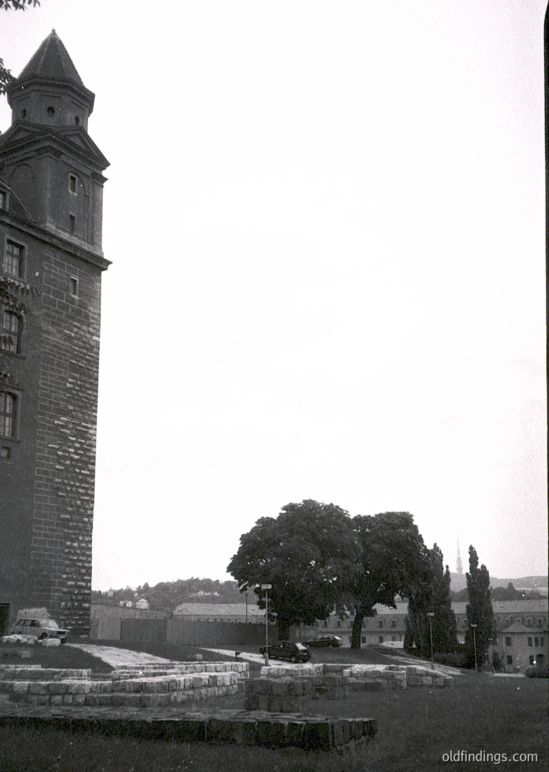 Historic stone tower with conical roof, likely part of a medieval fortress or watchtower. Surrounding ruins and low-lying buildings suggest a preserved or restored site. Overgrown tree in foreground adds natural contrast.