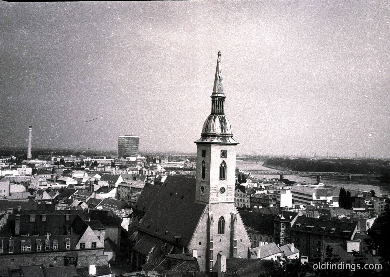 Aerial view of a historic European cityscape featuring a prominent **19th-century church tower** with a spire and clock, surrounded by dense, low-rise buildings. Industrial chimneys and modern high-rises contrast with traditional architecture, suggesting mid-20th century urban development. Likely **Central/Eastern Europe**.