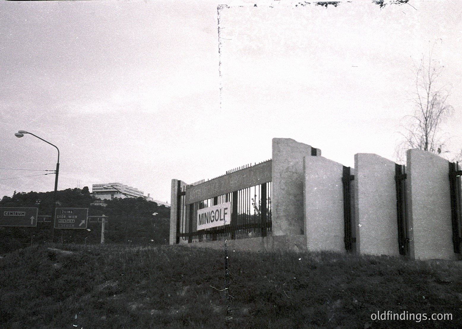 Vintage black-and-white photo of a concrete barrier with "MINGOLF" sign, likely a 1960s-70s roadside advertisement. Urban landscape includes directional road signs ("CENTRUM," "PLOVA DIVIZIA") and a distant building. Industrial-era road infrastructure with barbed-wire-topped concrete pillars.