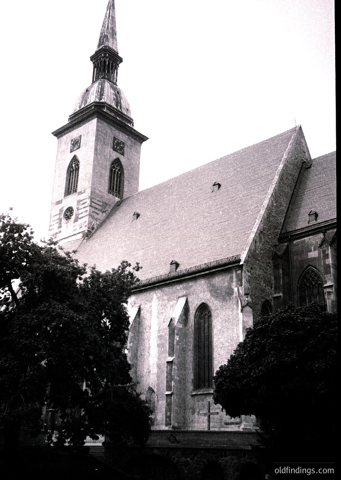 Historic church with Gothic Revival architecture—tall spire, pointed arches, and slate roof. Stone façade shows weathering, with a prominent cross on the gable. Surrounded by mature trees, suggesting a serene, possibly European setting.
