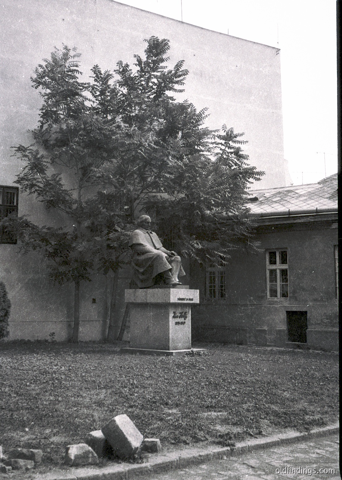 Seated bronze statue of a bearded figure in contemplative pose on a stone pedestal, set in a courtyard with gravel ground. Building with white-framed windows and a tree partially obscuring the statue. Likely mid-20th century Soviet-era monument.