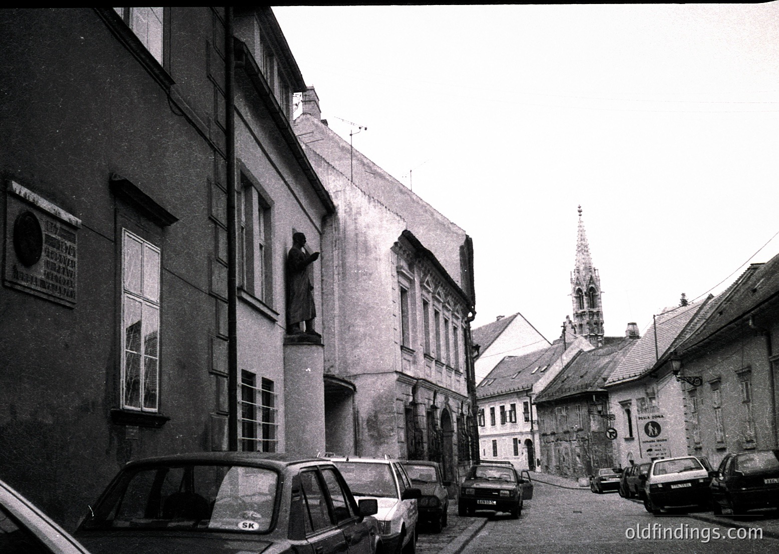 Vintage black-and-white street scene featuring Soviet-era cars parked along a narrow cobblestone alley. Prominent Soviet-era statue atop a building’s facade, flanked by older brick structures. Gothic-style church spire visible in background. Likely Eastern Europe, 1960s–1980s.