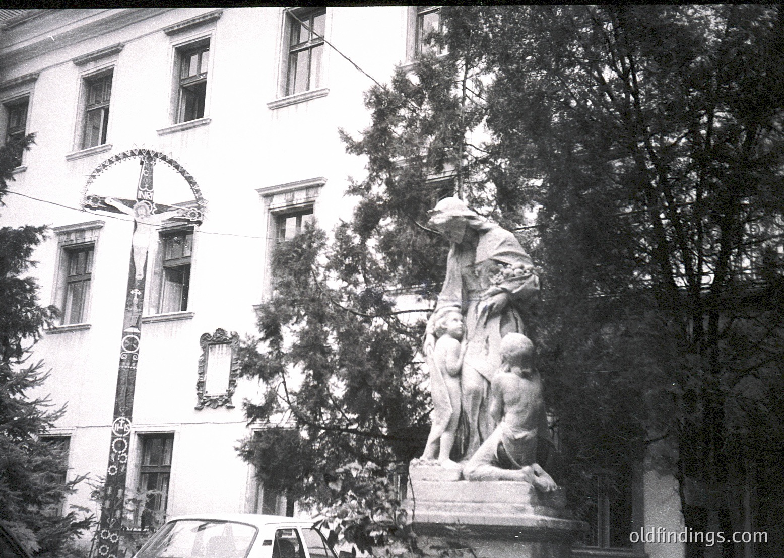 Neoclassical statue group depicting a seated woman cradling a child, flanked by a standing figure, set against a multi-story building with decorative wrought-iron balconies and arched windows. Mid-20th century urban setting, likely European.