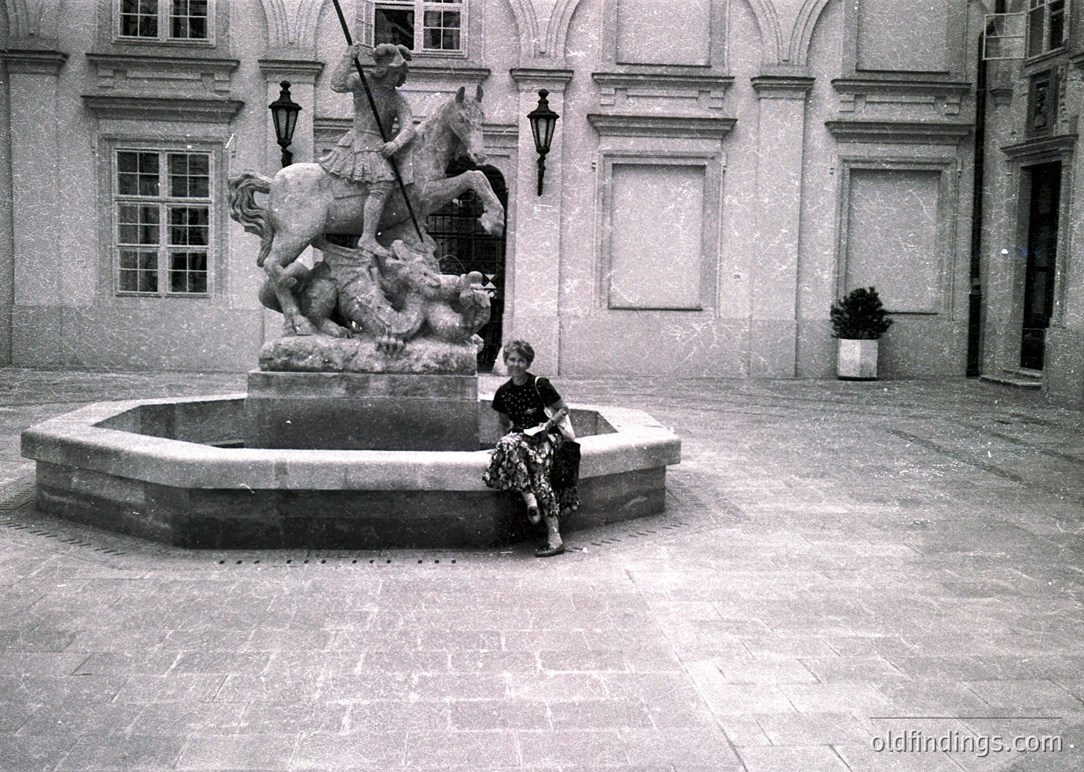 Neoclassical courtyard fountain featuring a knight on horseback slaying a dragon. Woman in 1960s-style dress sits on edge, holding a bag. Architectural details include arched windows and lanterns.