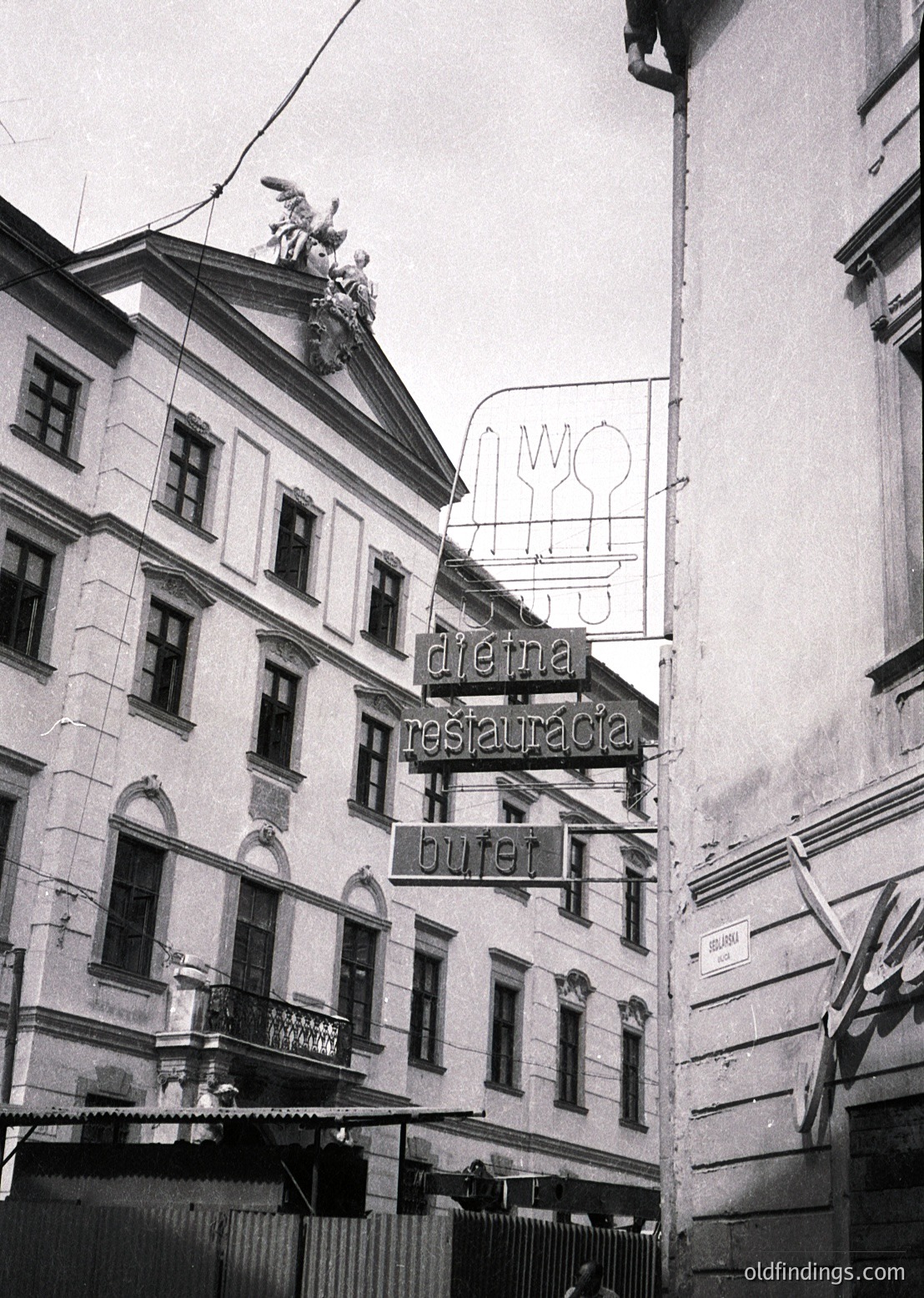 Neoclassical building corner with vintage signage: "Dieva Restauracia" (God’s Restaurant) in Cyrillic, featuring a stylized fork/knife logo. Architectural details include arched windows, decorative cornices, and a rooftop statue. Likely Eastern European, mid-20th century.