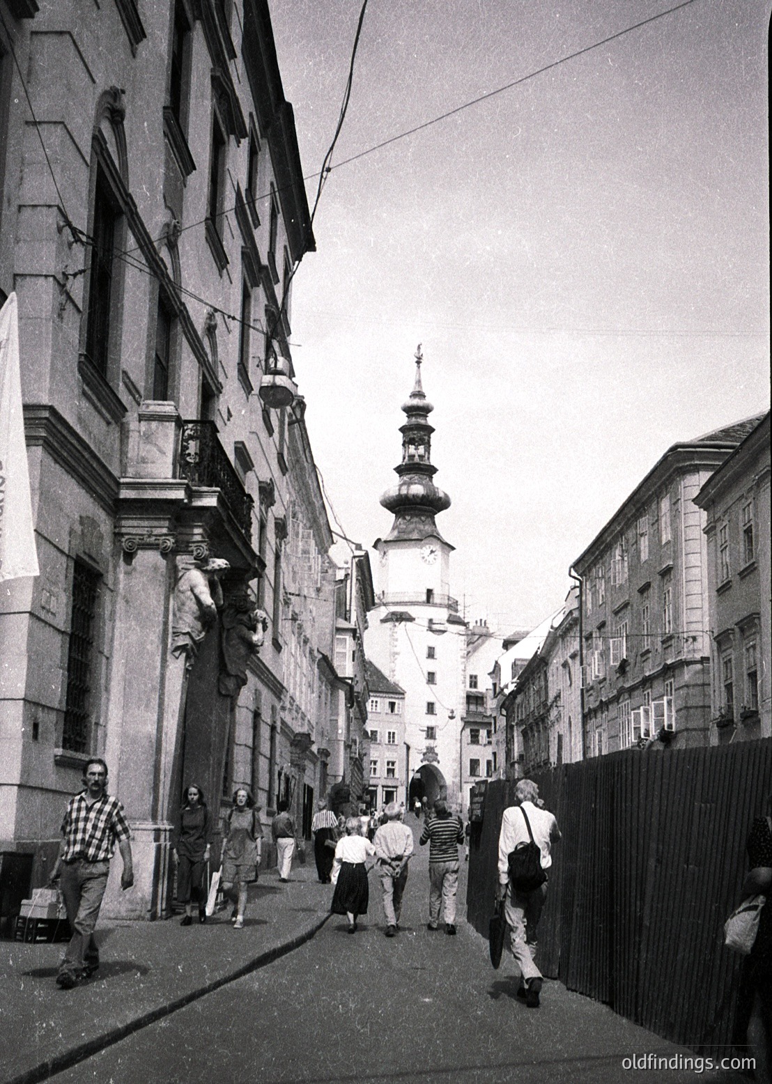 Narrow European street flanked by 19th-century stone buildings, leading to a Baroque-style church tower with onion dome. Crowd in 1960s-era clothing—checkered shirts, trousers, and hats—walking toward the tower. Urban architecture reflects Soviet-era urban planning in a Central/Eastern European city.