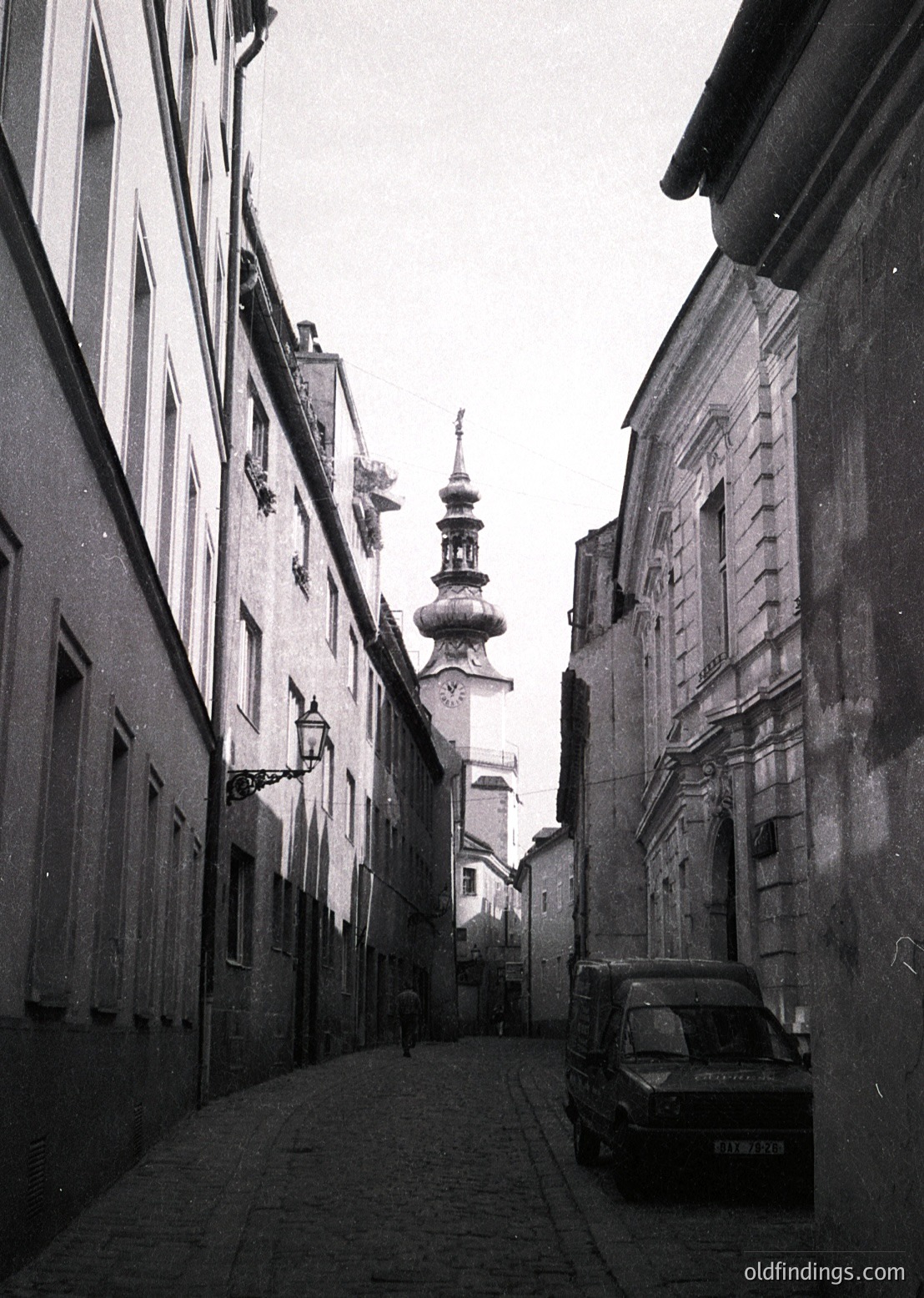Narrow cobblestone alley flanked by historic European buildings, leading to a domed church tower. Mid-20th century vintage car parked on right. Architectural details include ornate facades and wrought-iron street lamps. Likely Eastern European setting.