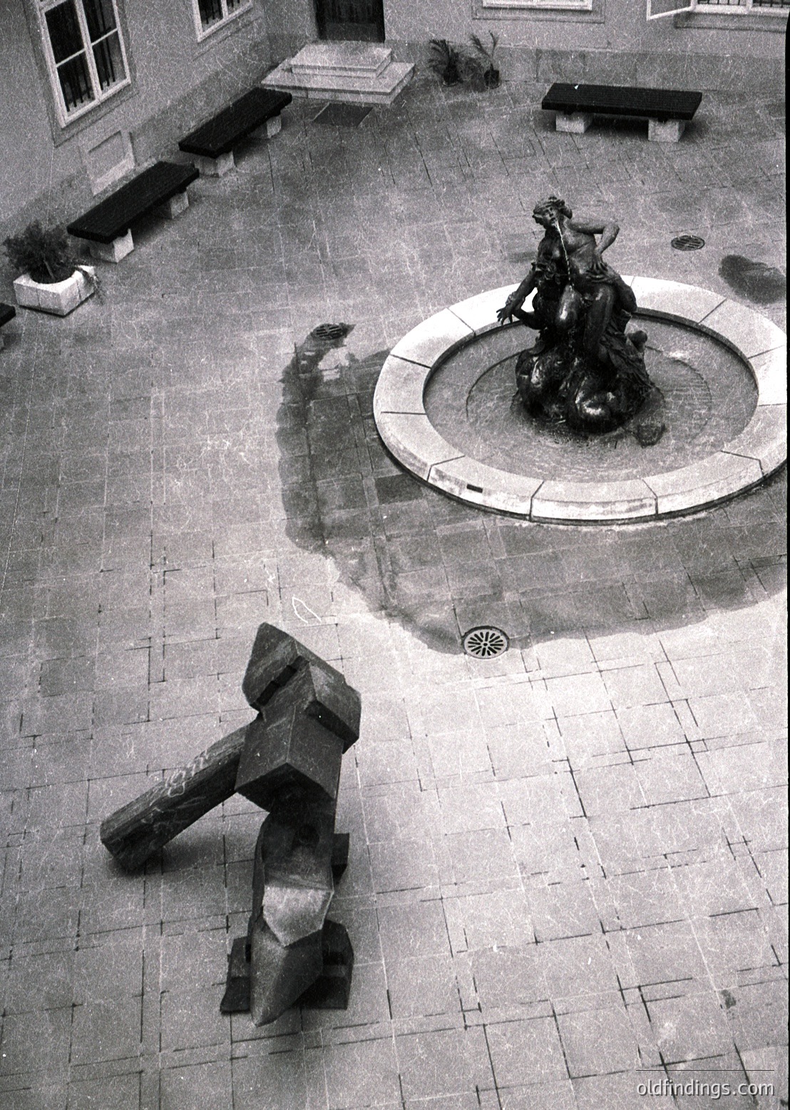 Aerial view of a courtyard featuring a classical fountain with a seated female figure holding a bowl, surrounded by a circular stone basin. Adjacent to it, an abstract concrete sculpture resembling a bent arm or abstract human form. Surrounding architecture includes stone walls, benches, and potted plants.